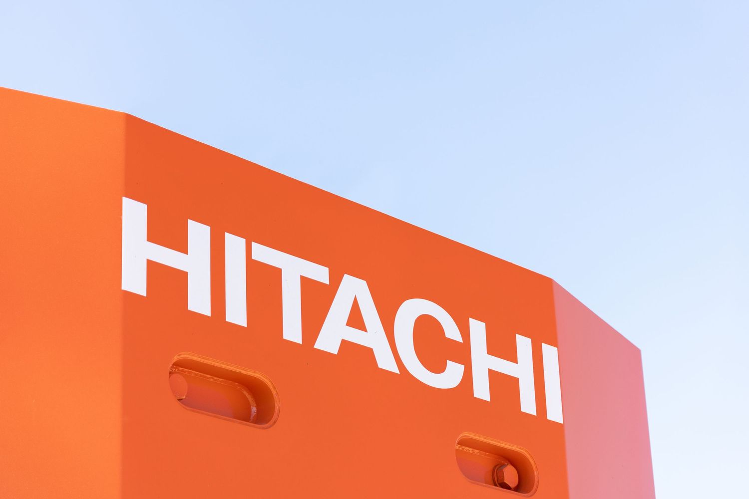 Large white Hitachi corporate logo on orange surface against blue sky background.