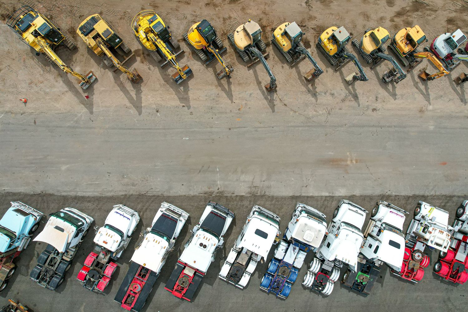 Aerial view of construction vehicles and trucks parked in neat rows on dusty ground with distinct shadows.