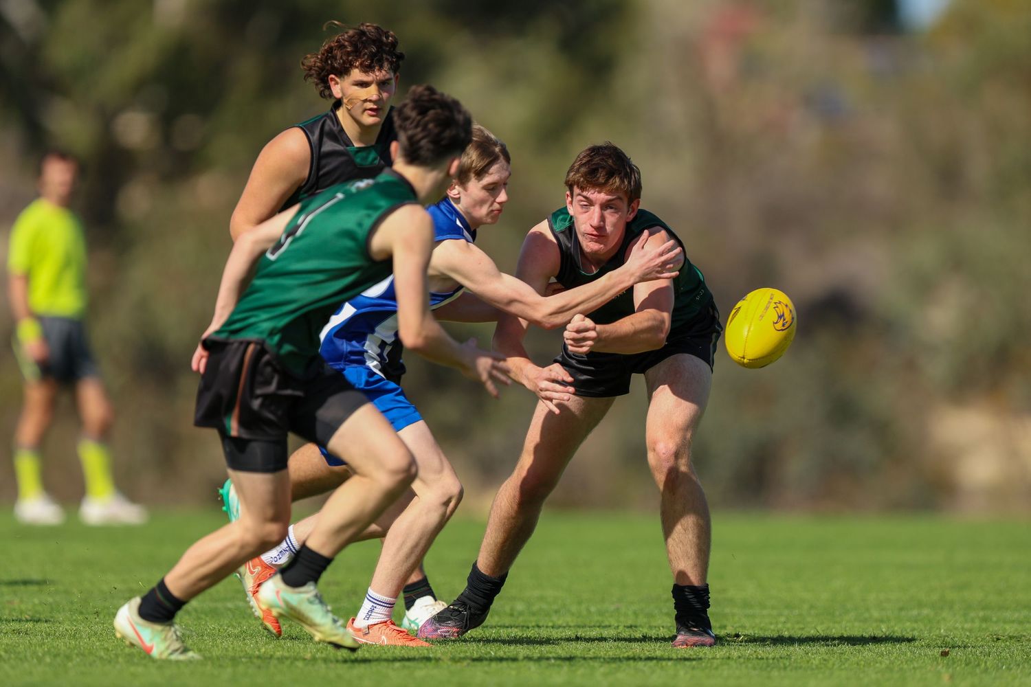 Athletes dive and reach for the ball during a competitive Australian Rules Football game on a sunny field.