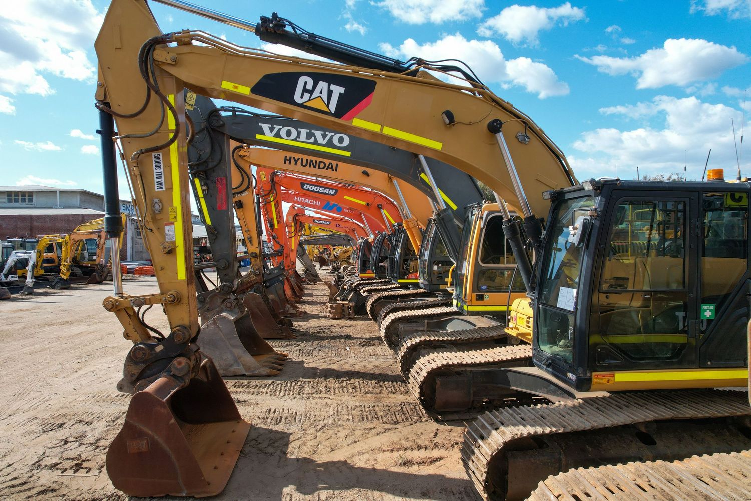 Line of yellow CAT and Volvo excavators parked in a row at a construction equipment yard under blue sky.