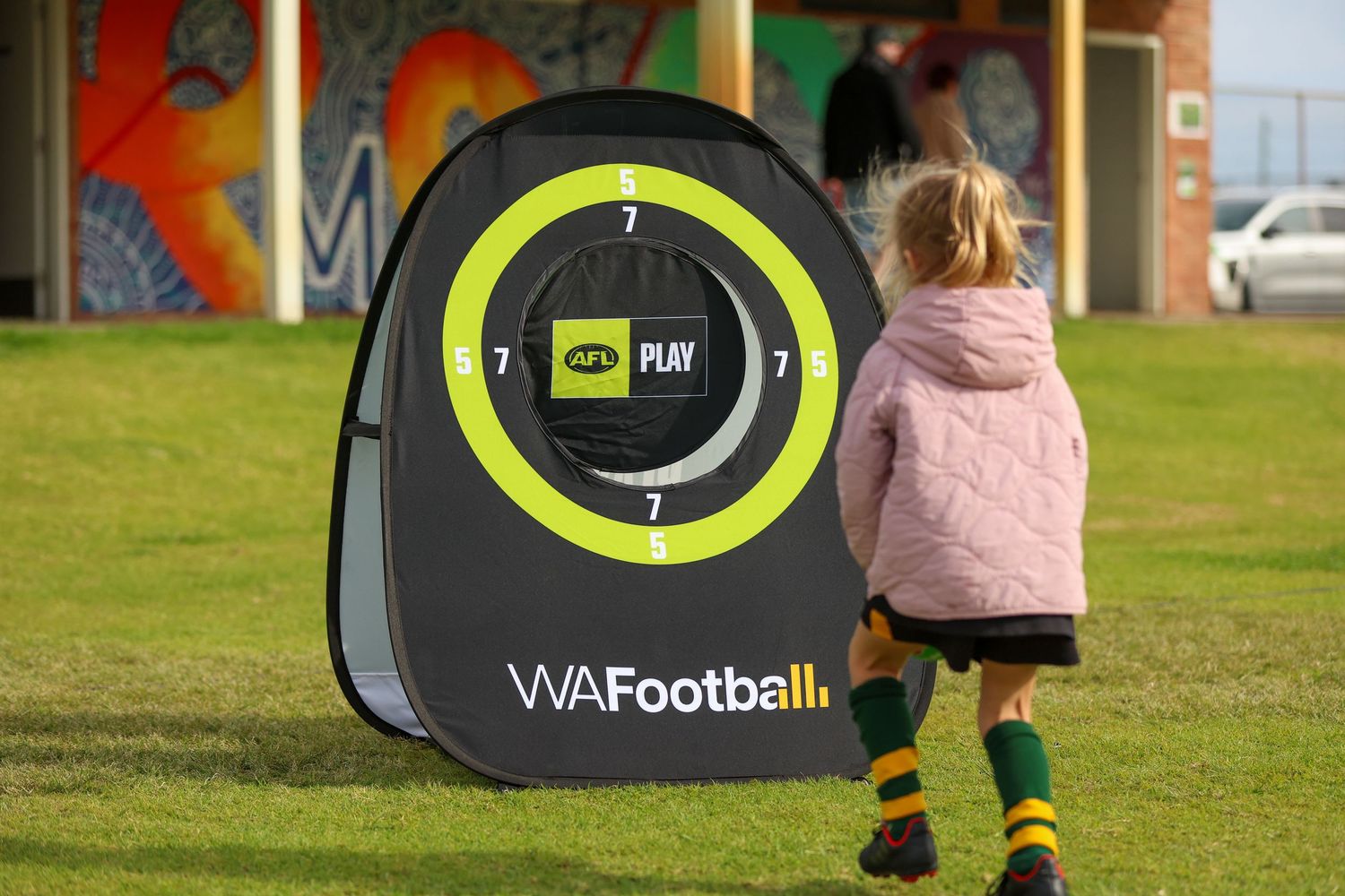 A child in a pink jacket and yellow socks stands in front of a WA Football training target on a grassy field.