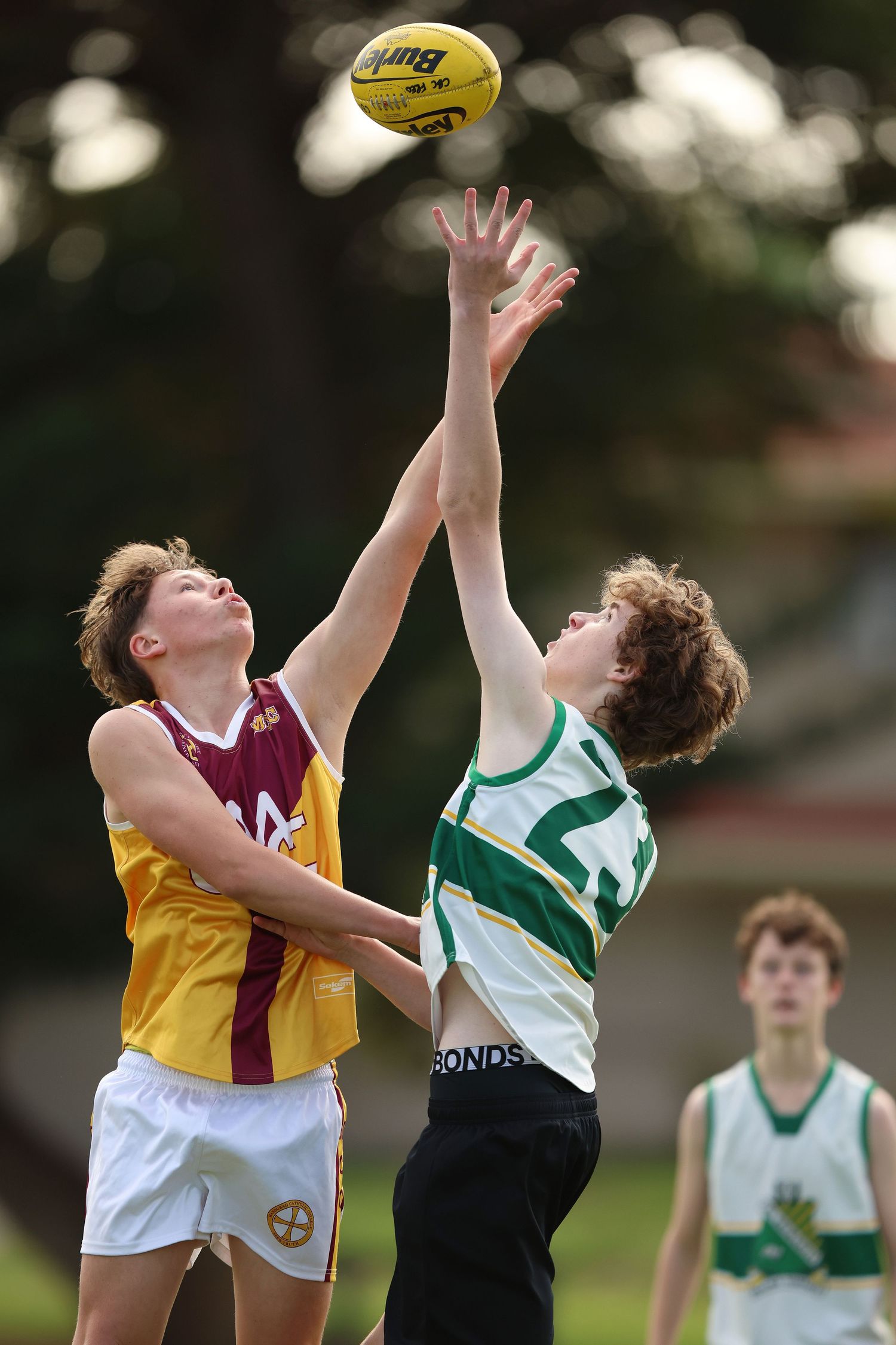 Two young Australian Rules Football players leap high to compete for the ball during a match, with another player watching.