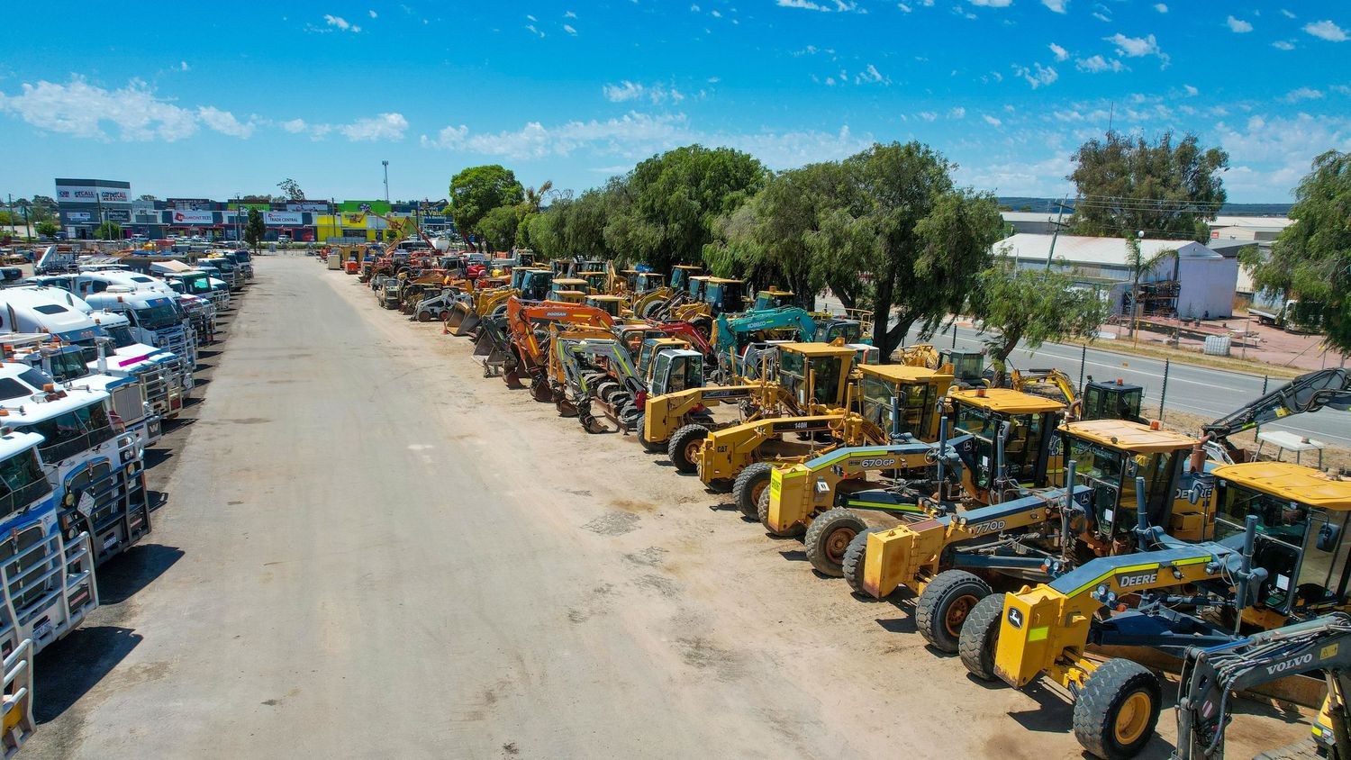 Long row of yellow construction equipment and vehicles parked on a dirt road at an industrial auction yard.