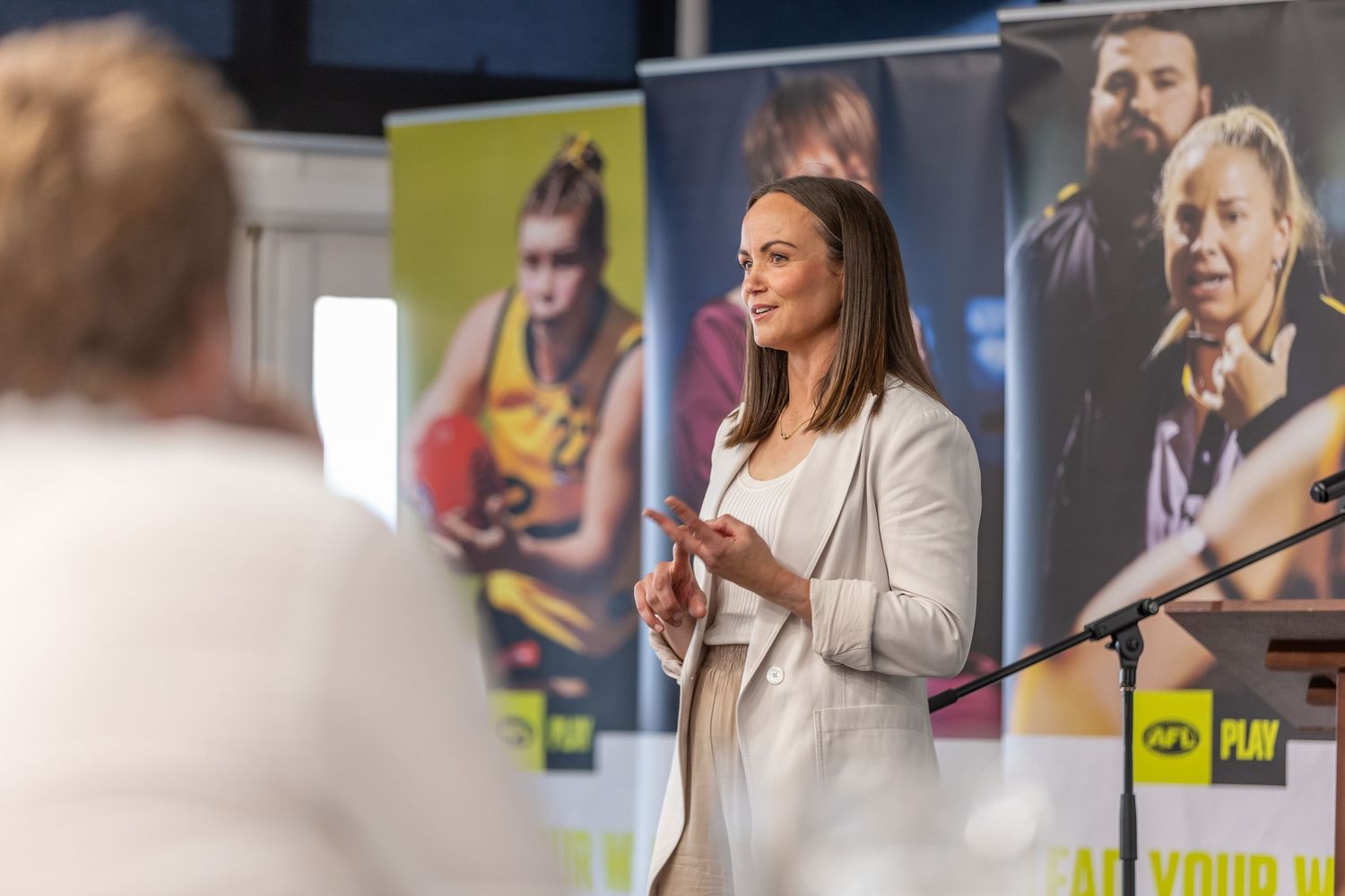 A presenter speaks on stage with sports posters displayed in the background during a conference.