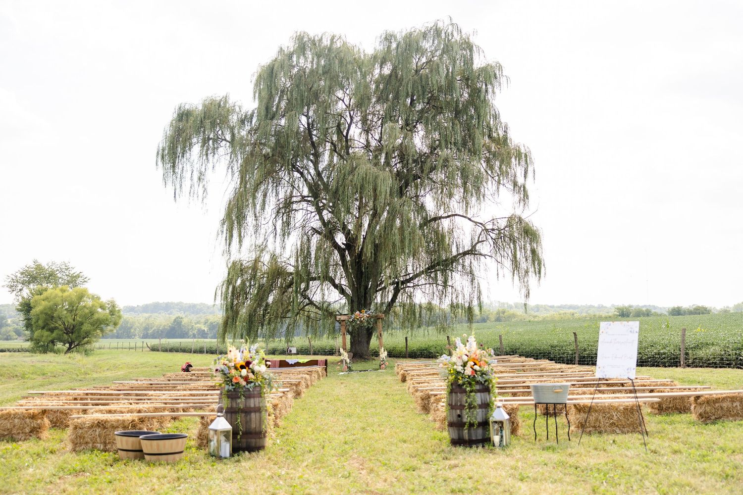 Outdoor farm wedding ceremony setup with hay bale seating, barrel florals, and weeping willow tree backdrop.