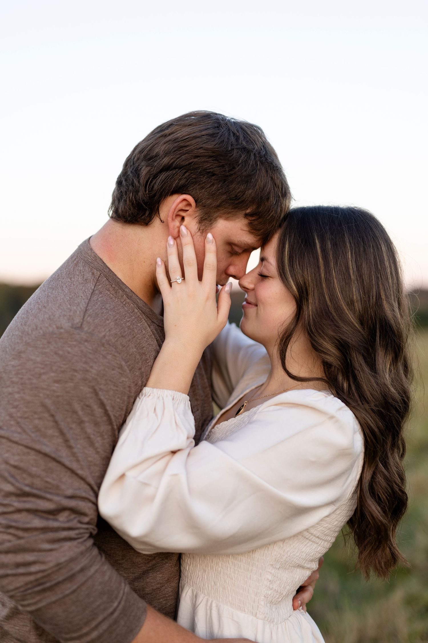 A couple shares a romantic forehead touch at sunset, woman in white dress with engagement ring visible.