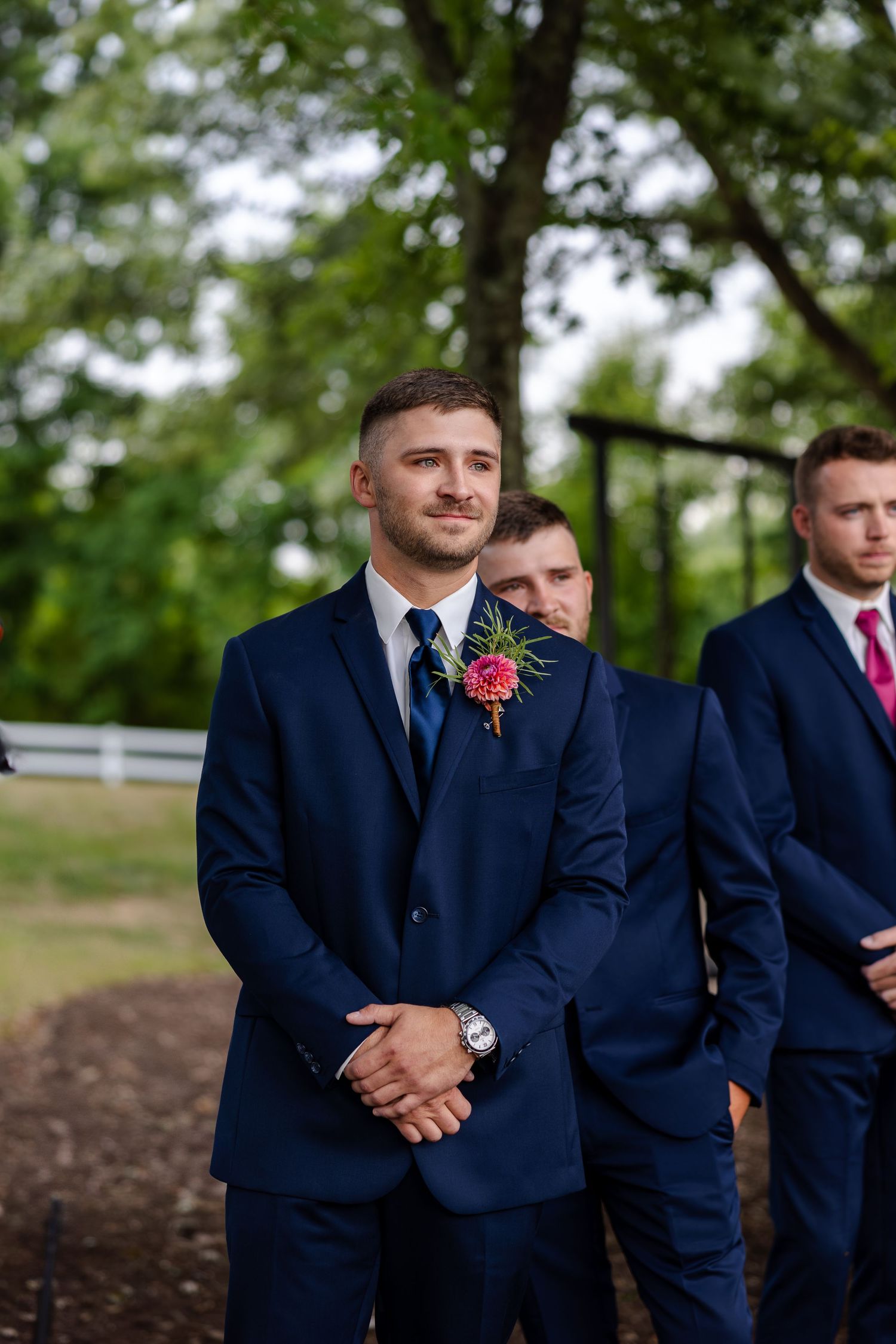Groom in navy suit with pink boutonniere stands with groomsmen at outdoor wedding ceremony.