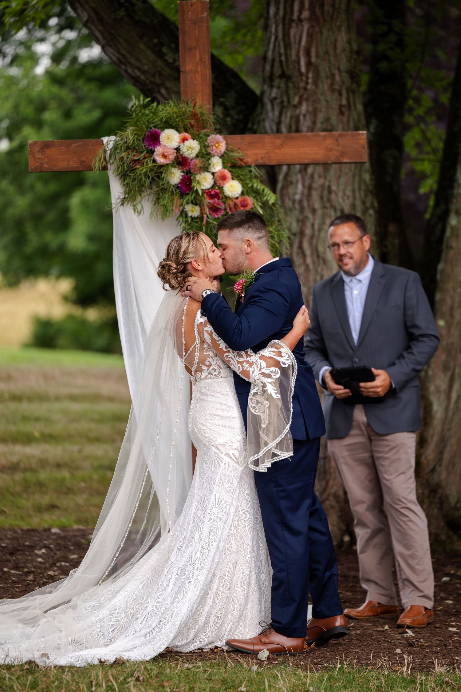Bride and groom share first kiss at outdoor ceremony before floral-decorated wooden cross.