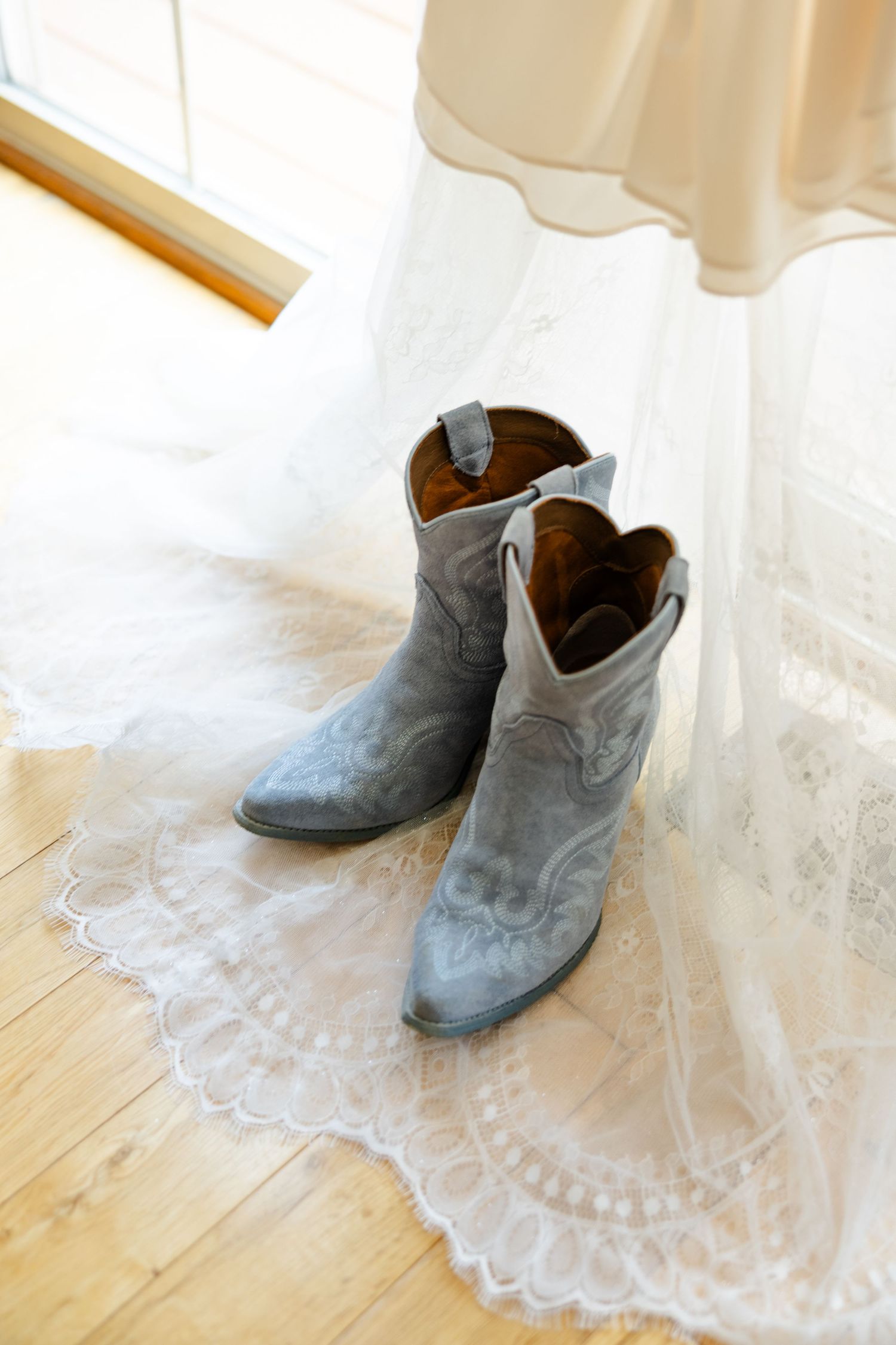 Blue suede cowboy boots sitting beneath a lace wedding dress hem on a hardwood floor near a window.