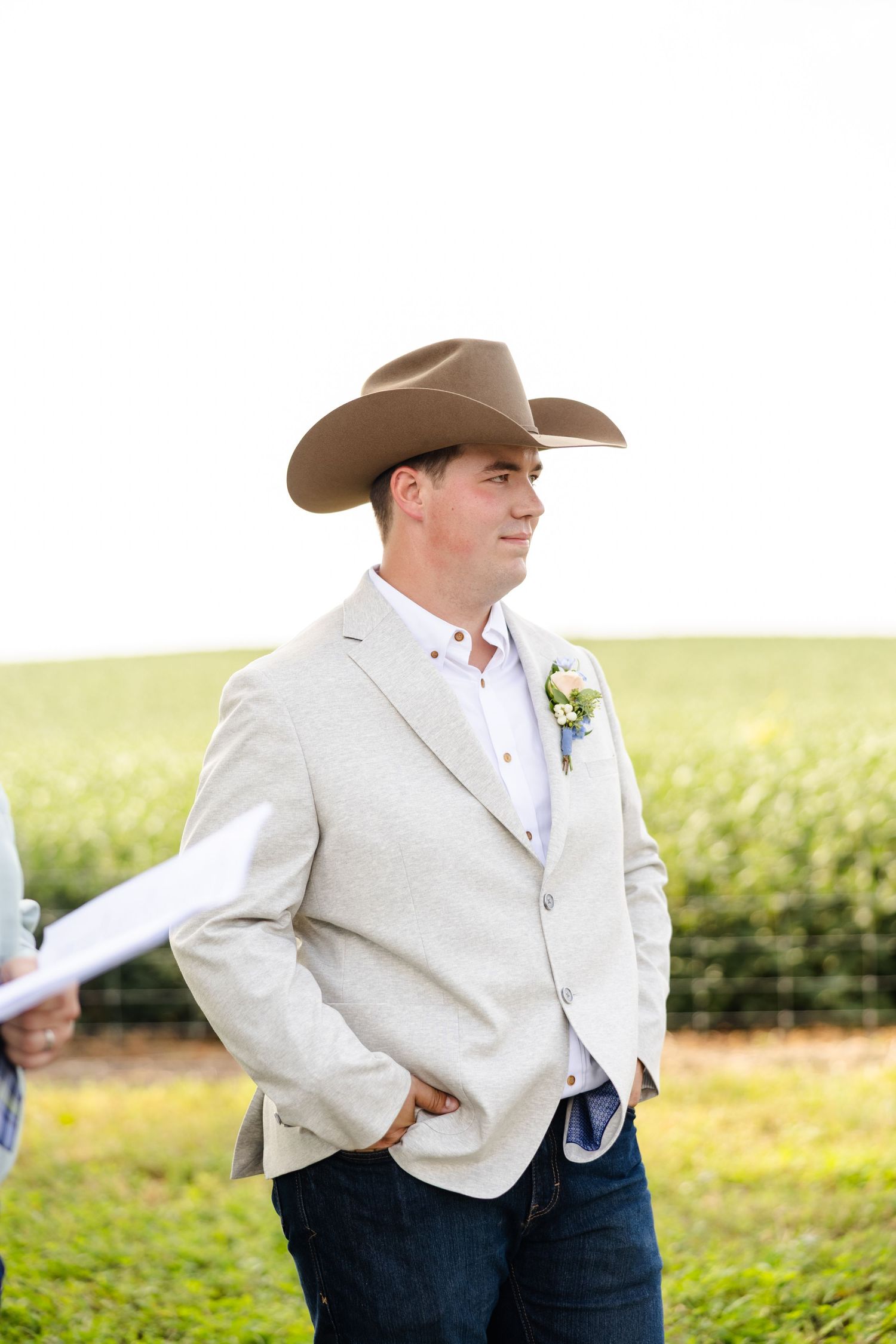 Handsome groom wearing a cowboy hat and tan blazer with boutonniere standing at outdoor farm wedding ceremony.