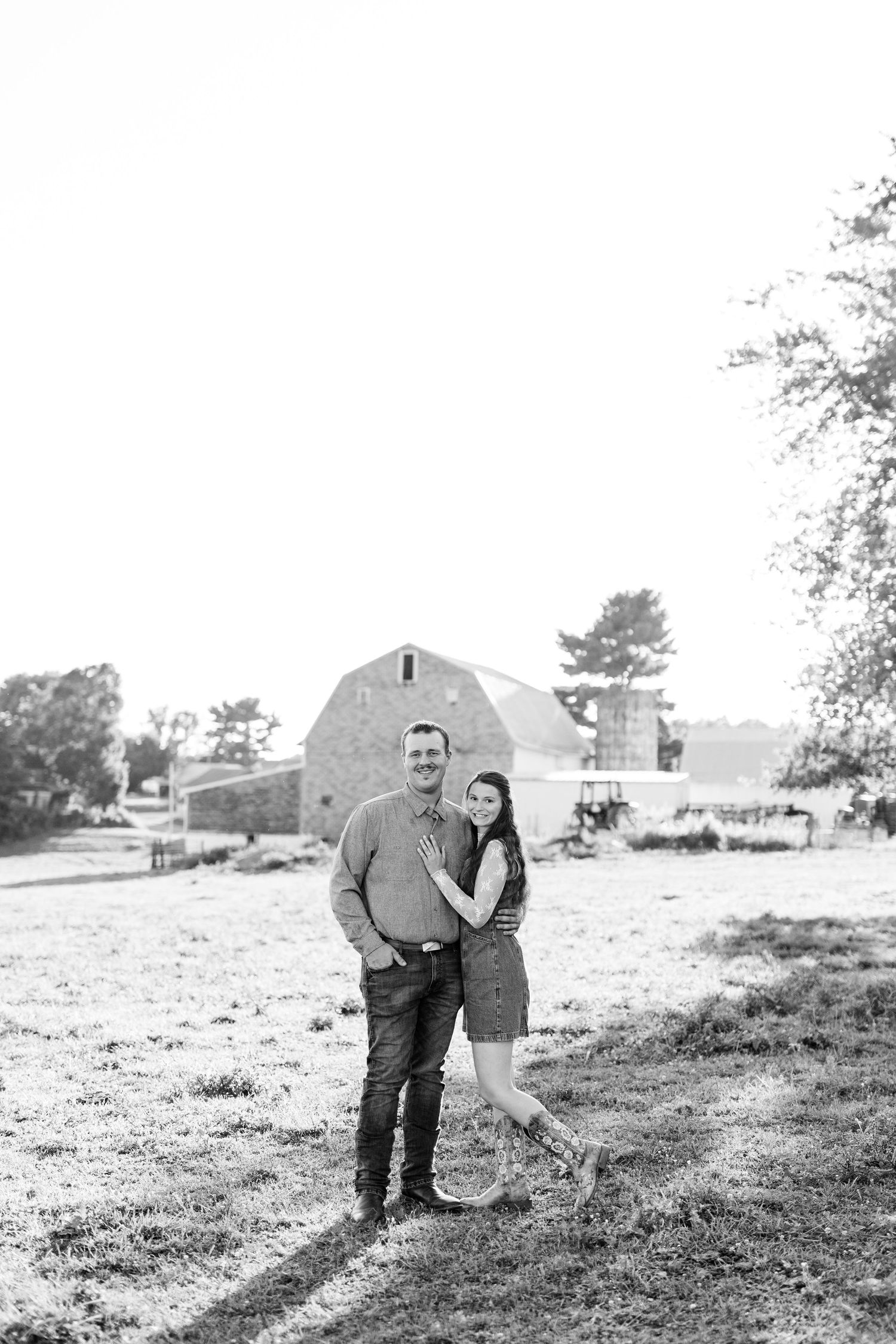 Black and white photo of a couple posing together in front of a rustic barn on a farm.