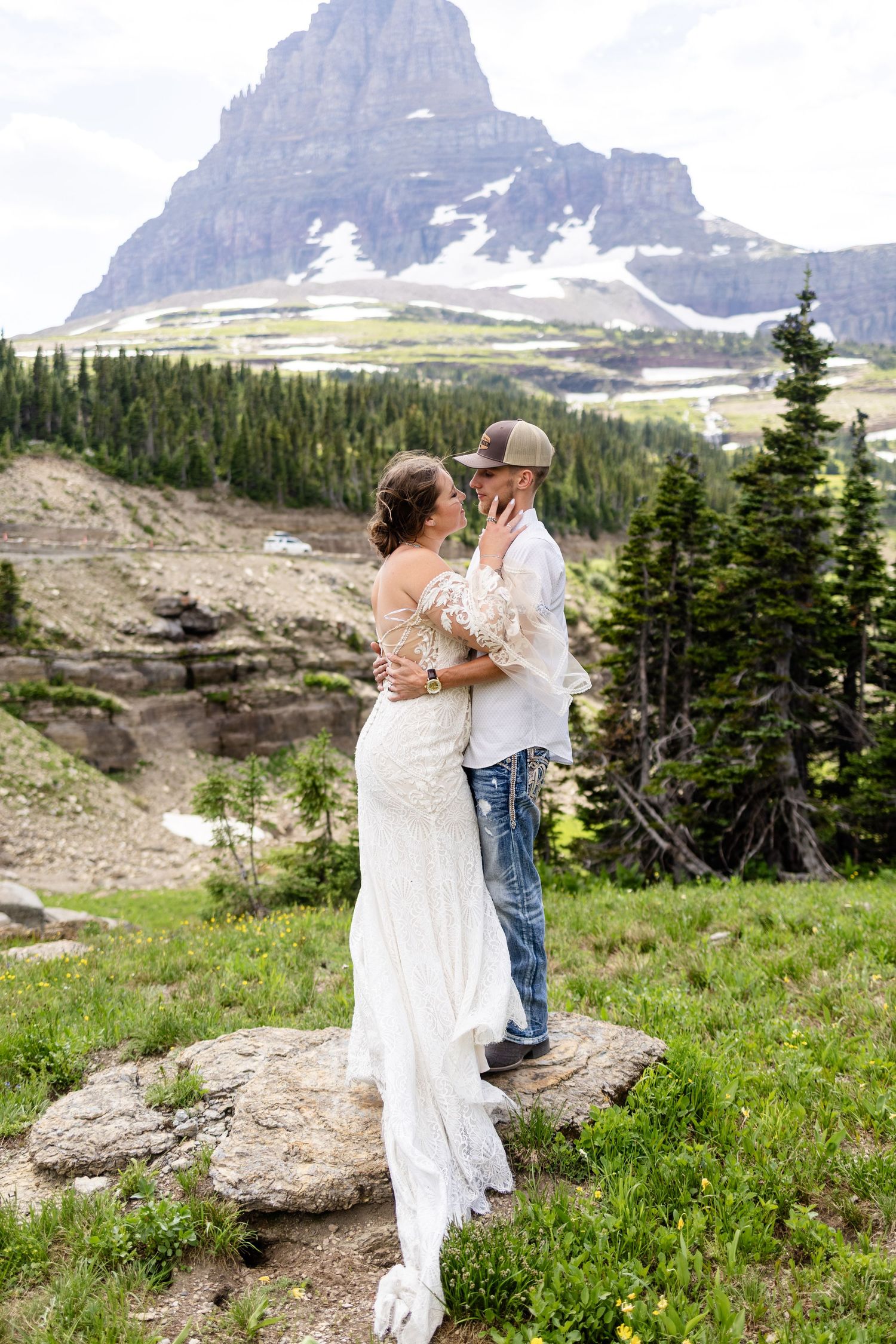 Bride and groom embracing on a rock with stunning mountain peak and alpine meadow scenery behind them.