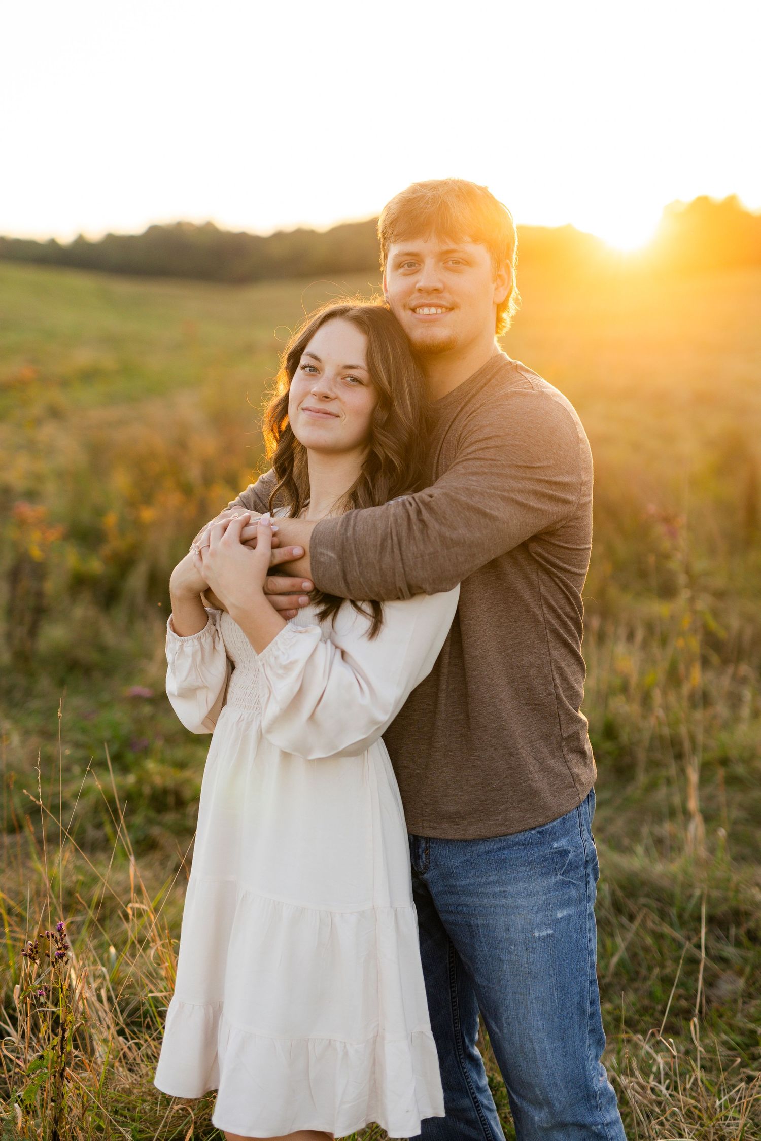 Couple embracing in a golden sunset field, woman in white dress, man in brown shirt and jeans, romantic outdoor portrait.