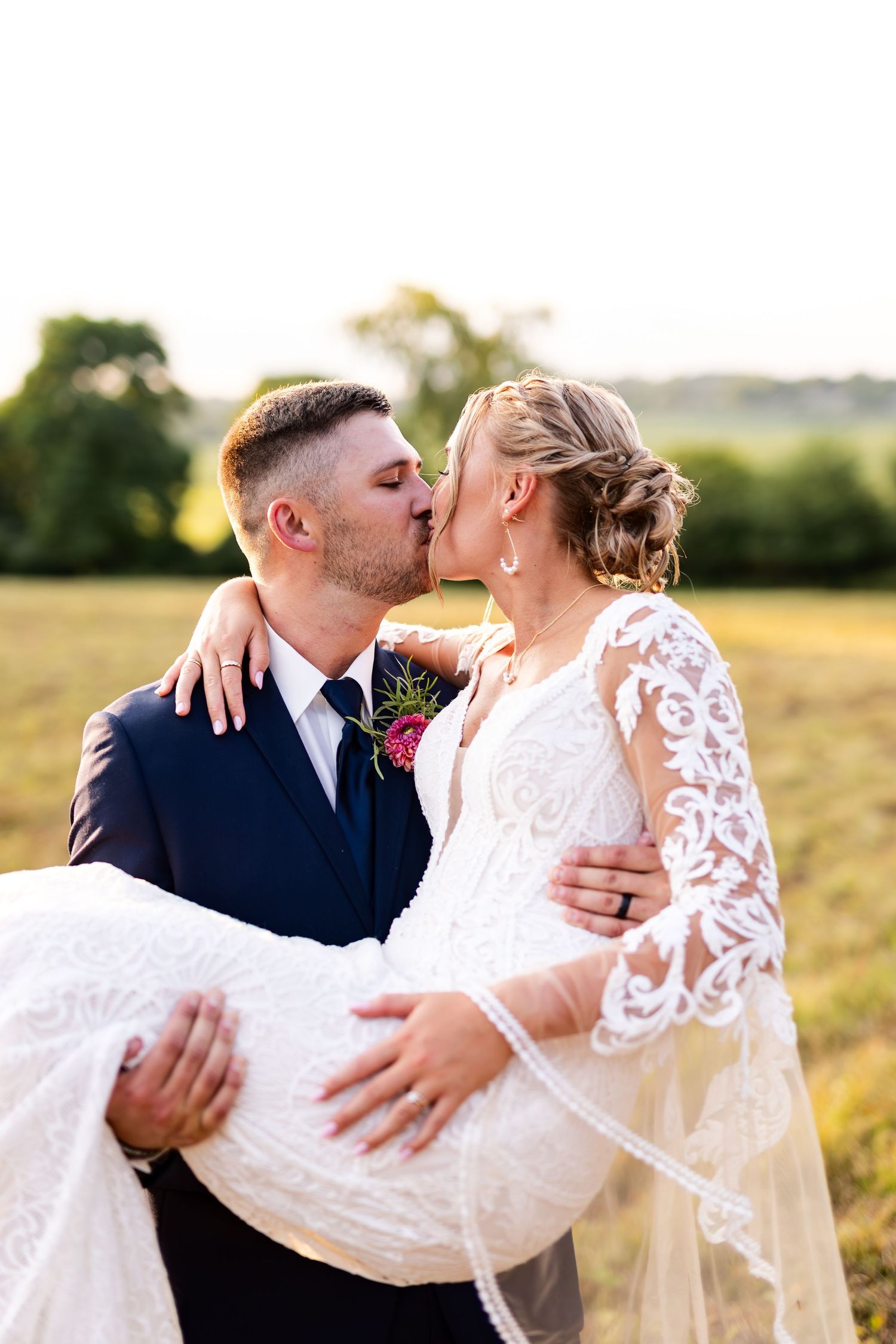 Groom in navy suit kisses bride while carrying her in a sunlit outdoor field setting.