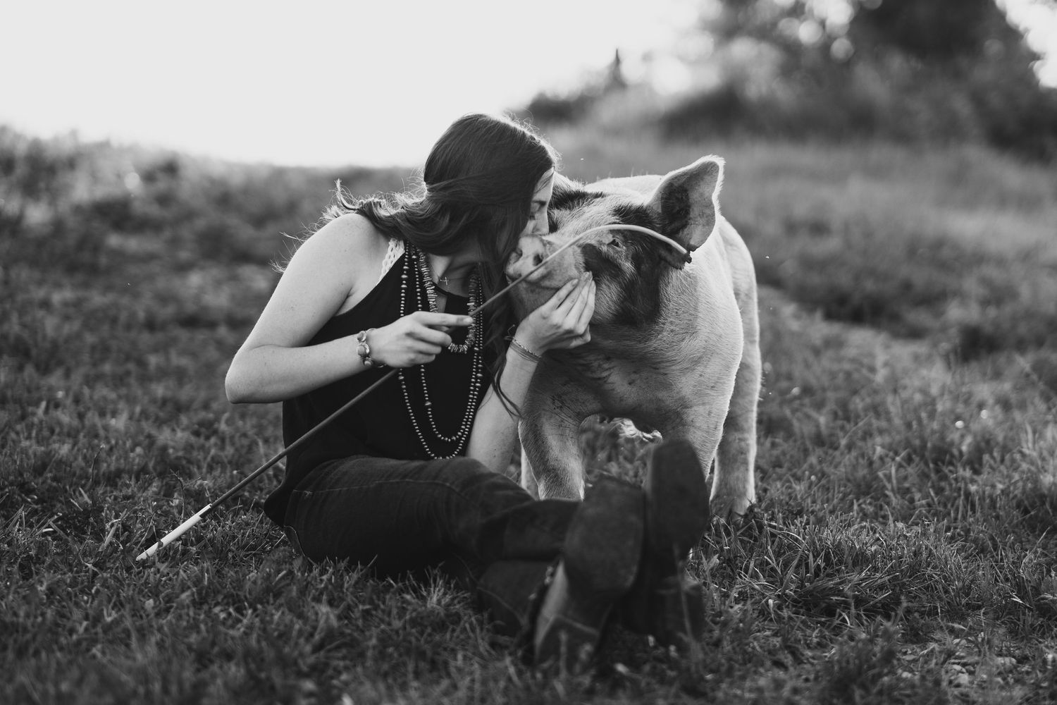 Black and white photo of a woman sitting on grass, affectionately nuzzling a pig outdoors.