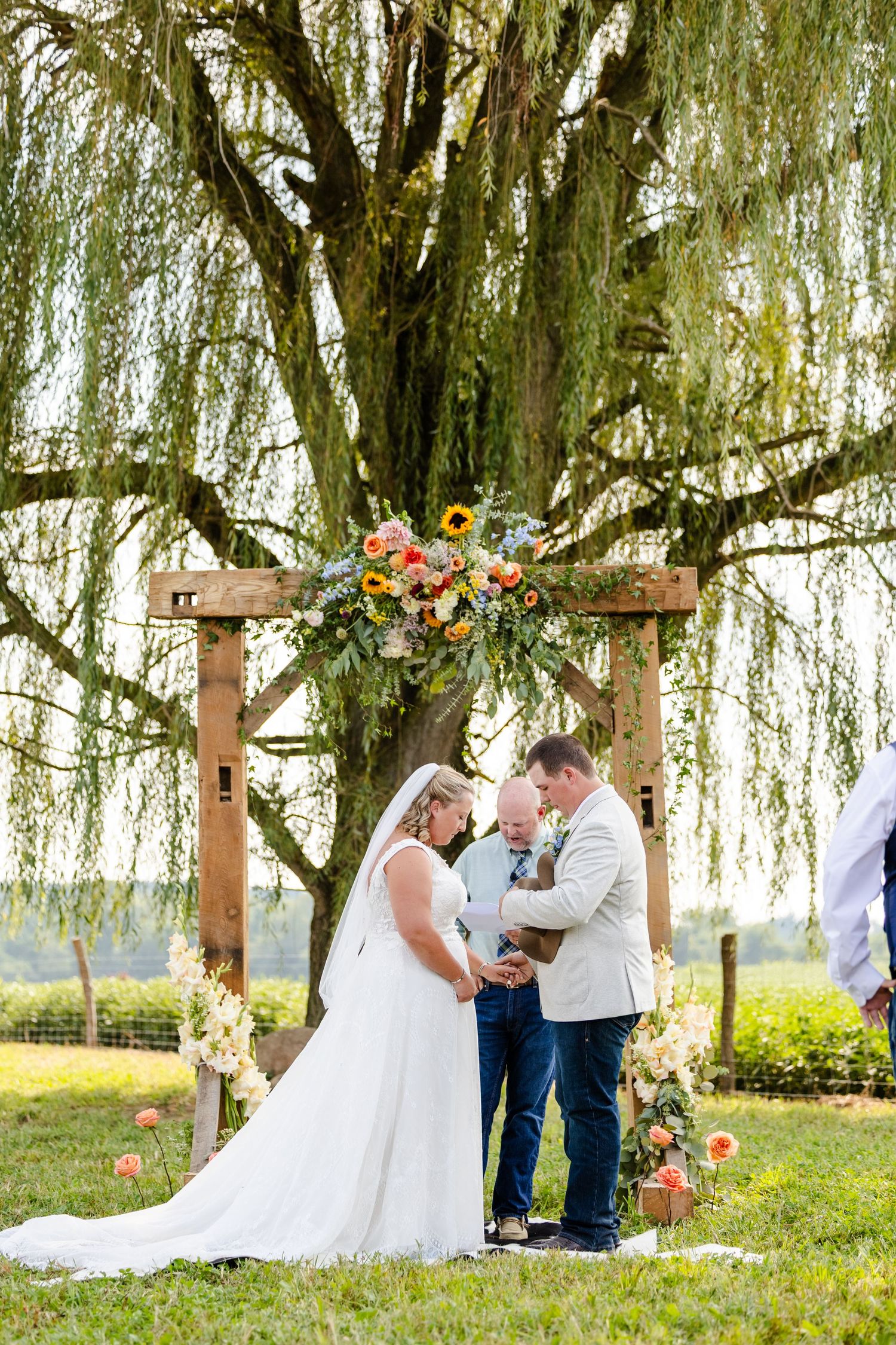 Bride and groom share an intimate moment under a floral wooden arch beside a weeping willow at an outdoor wedding.
