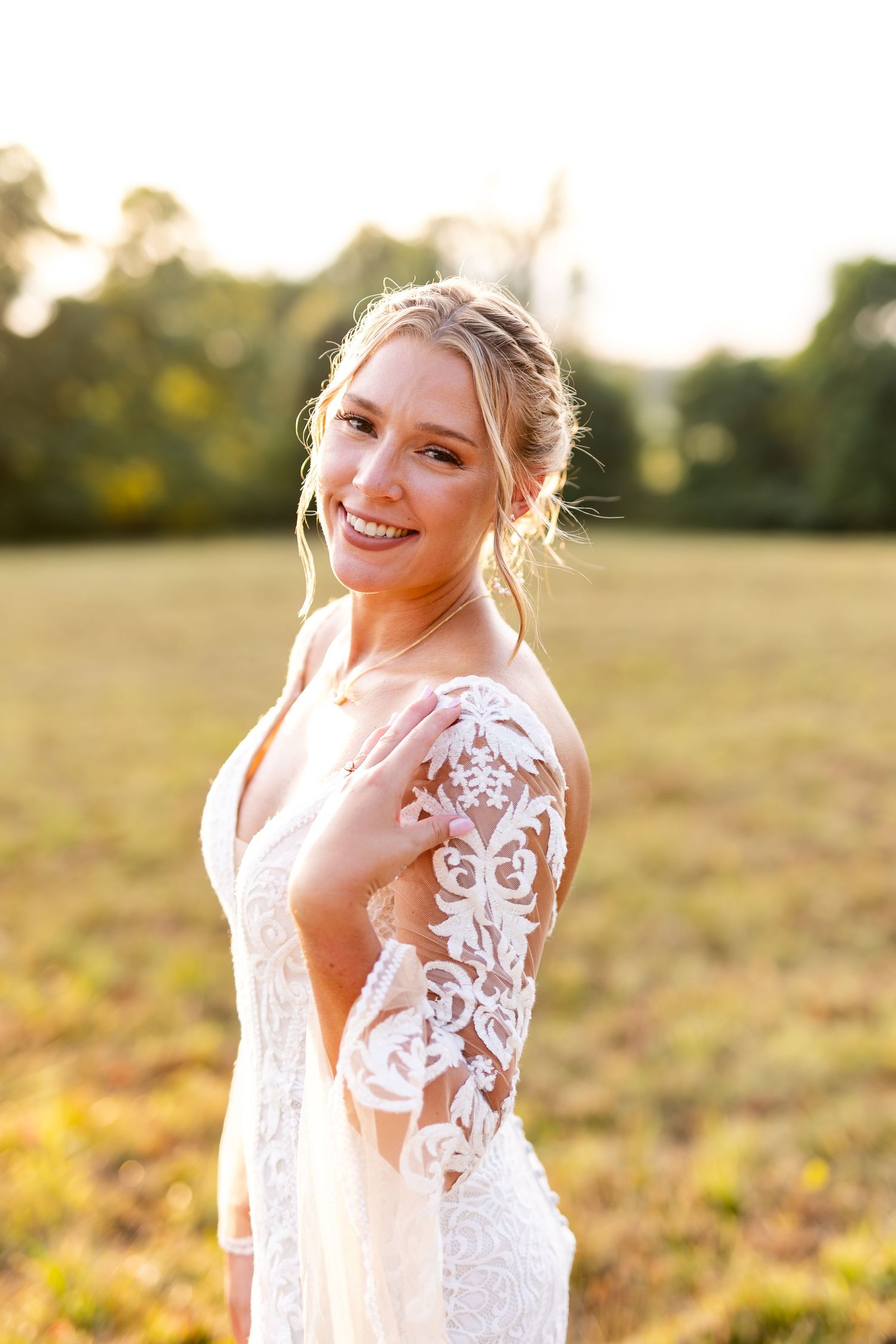 Smiling bride in lace long-sleeve wedding dress poses in golden sunlit field at sunset.