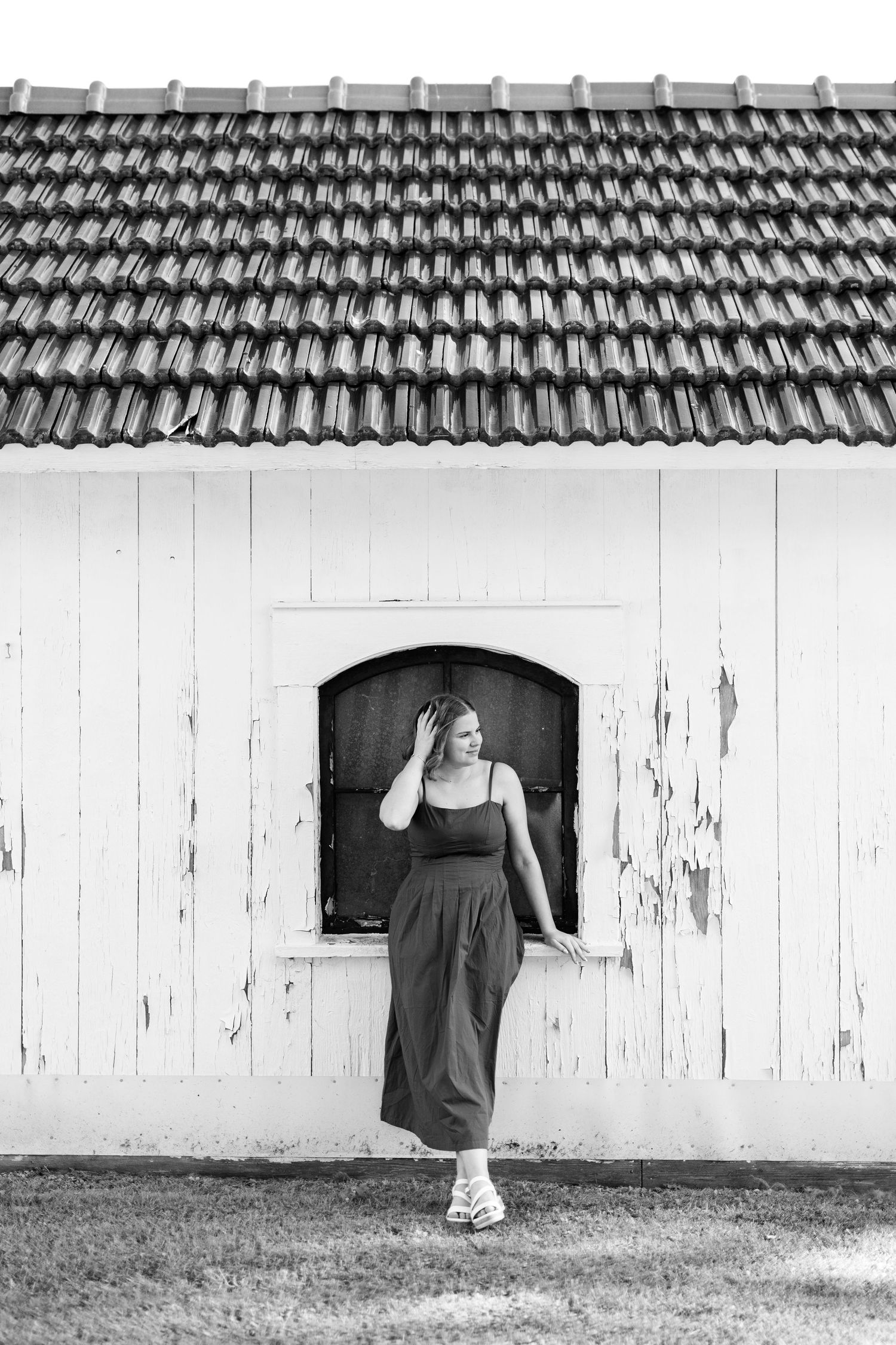 Black and white photo of a woman in a maxi dress posing by a rustic white barn with arched window and tiled roof.