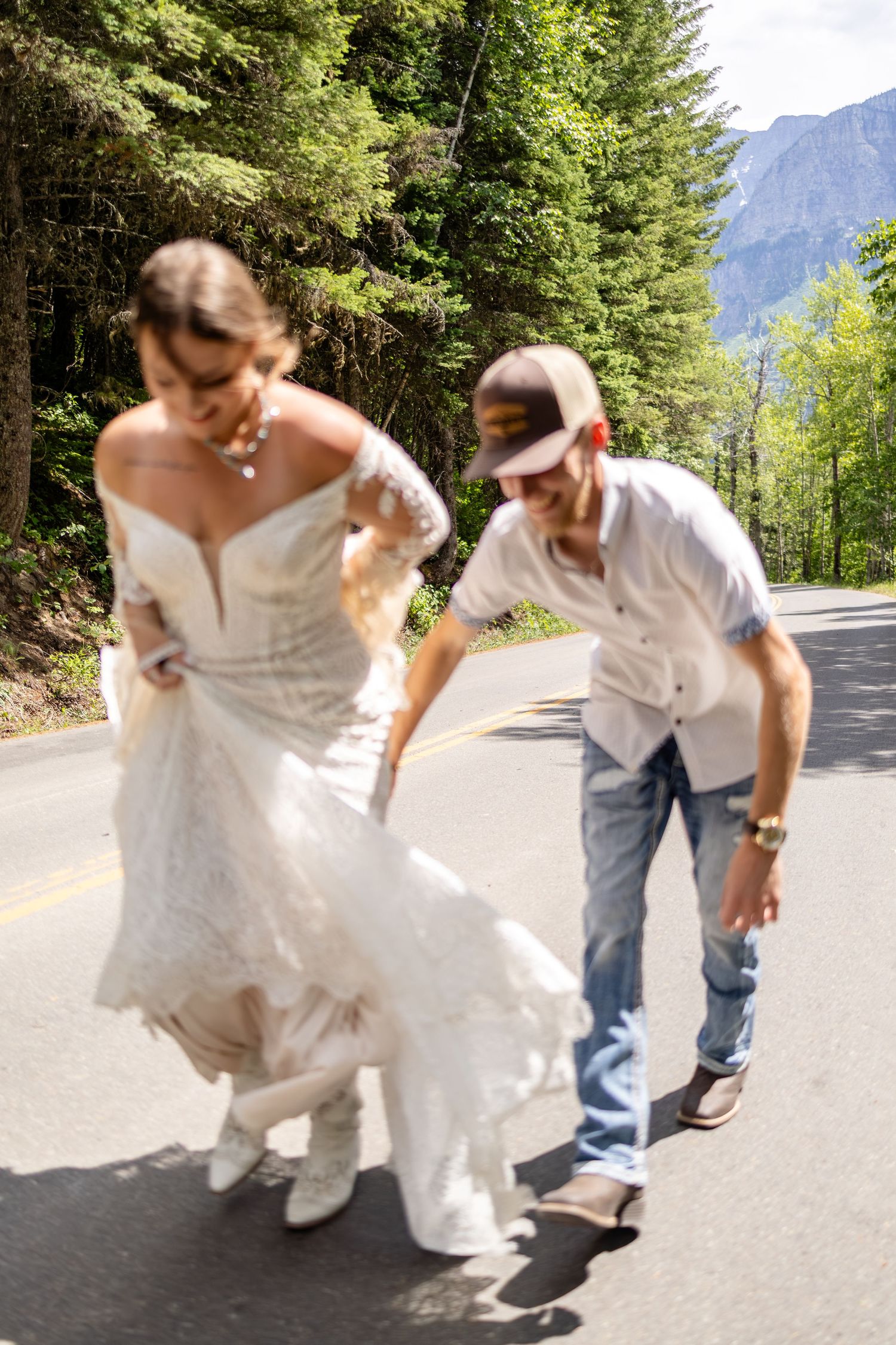 Bride in white lace gown and groom in jeans adjust her dress on a scenic mountain road surrounded by trees.