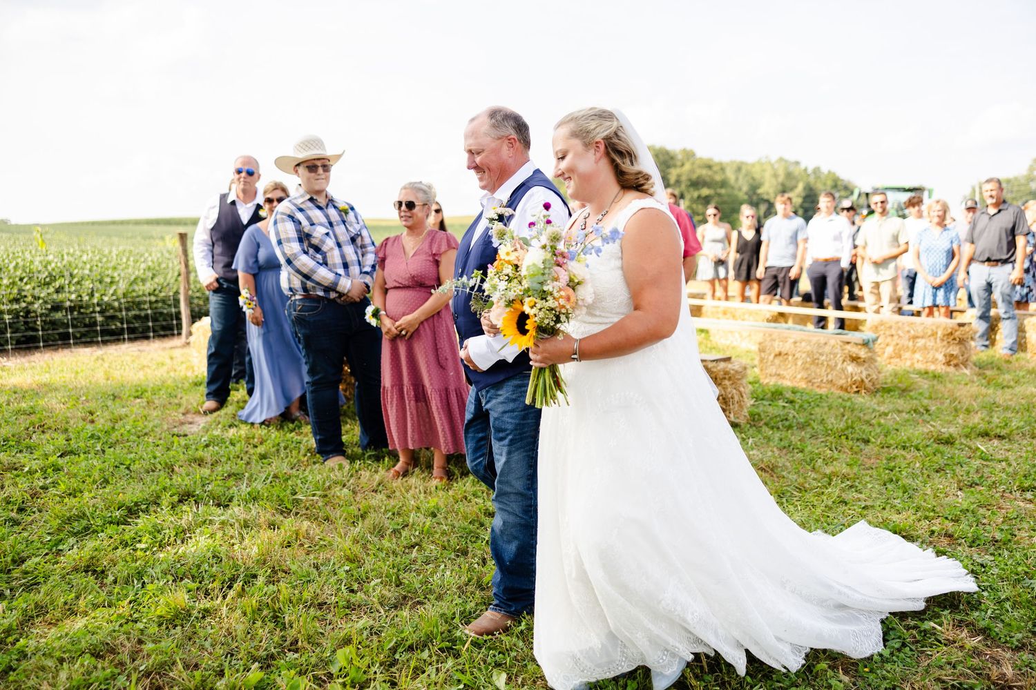Bride in white lace gown holding sunflower bouquet walking the aisle with escort at outdoor farm wedding.