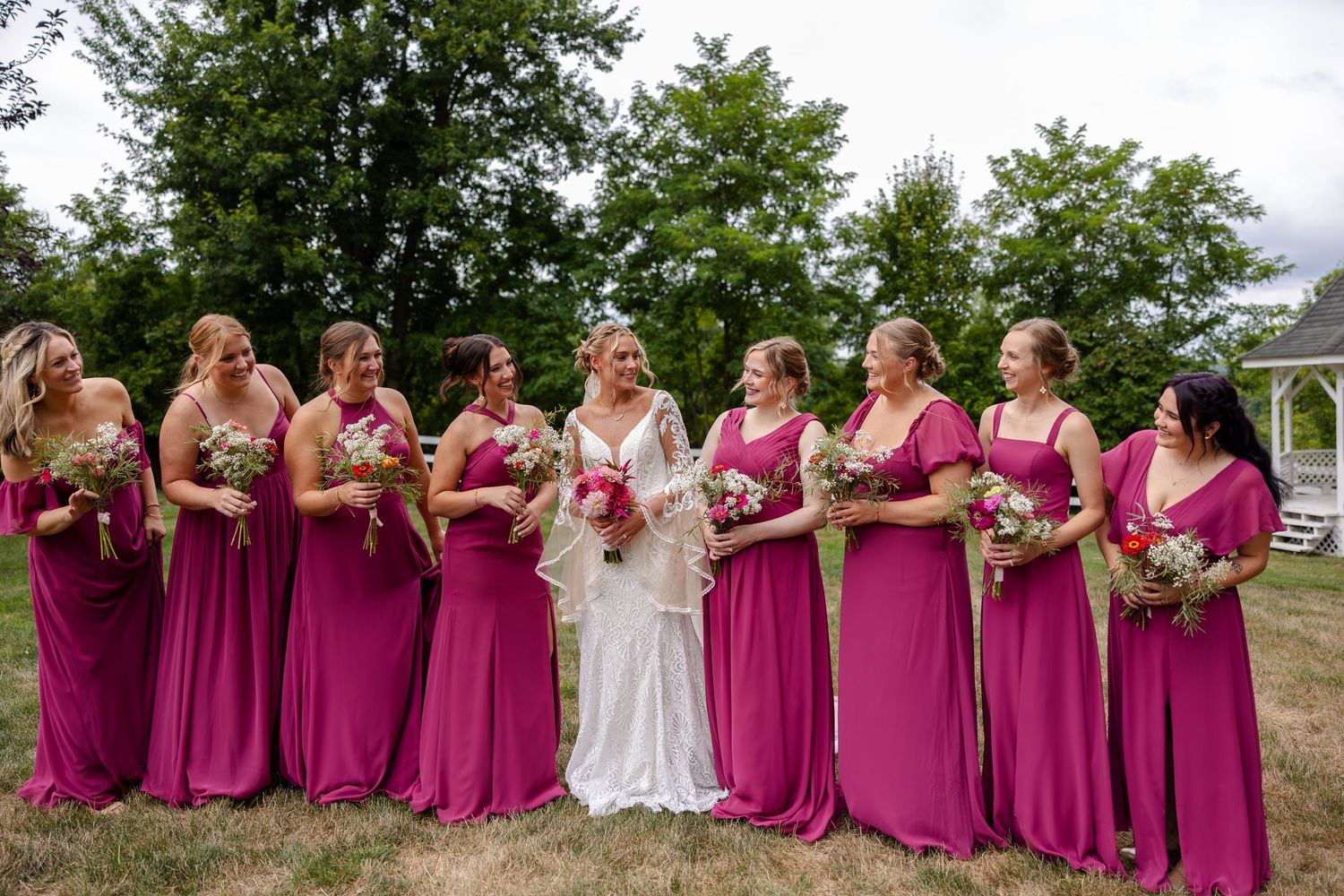 Bride and bridesmaids in pink dresses laughing together while holding floral bouquets on a green lawn.