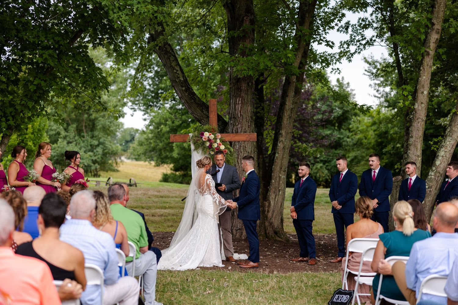 Couple exchanges vows at outdoor wedding ceremony beneath large tree with guests seated nearby.