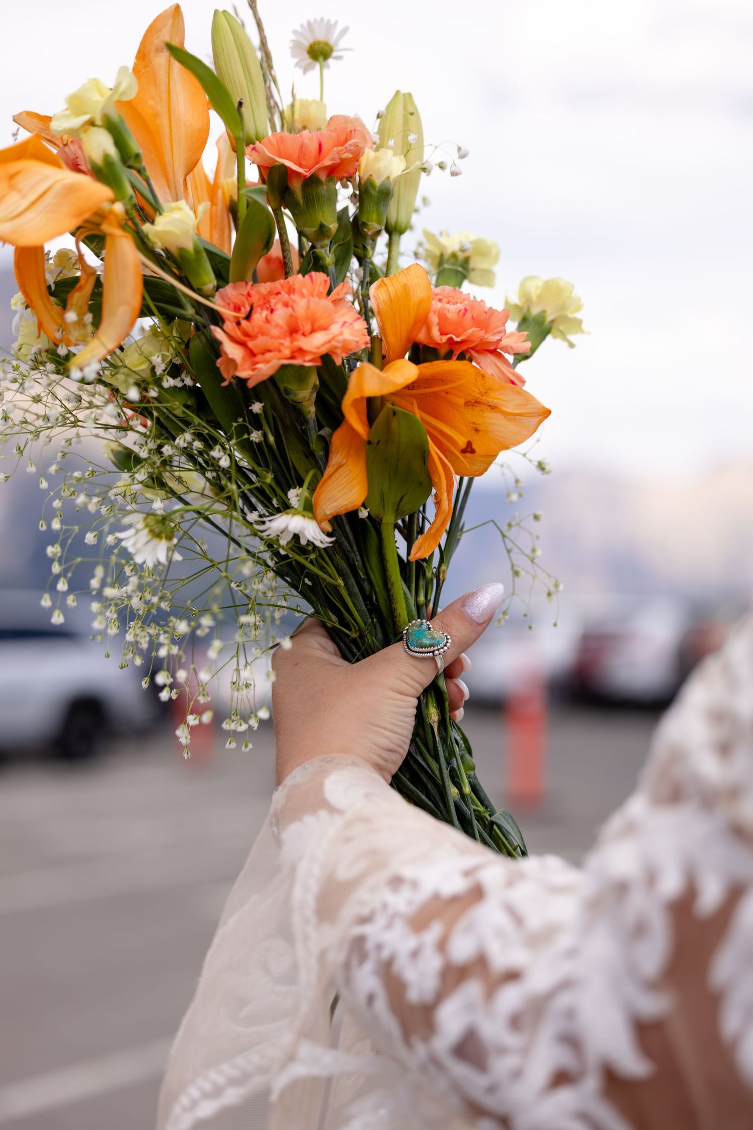 Bride holding a vibrant orange bouquet of lilies, carnations, and daisies with baby's breath, wearing a lace wedding dress.