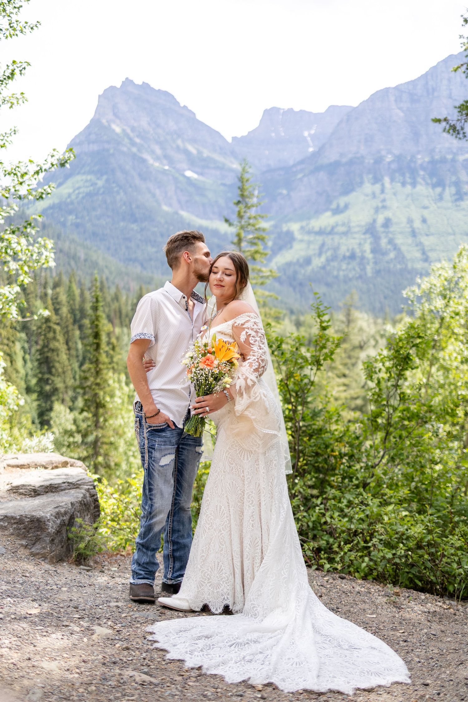 Couple shares a kiss in wedding attire with mountain and forest backdrop, bride holding colorful bouquet.