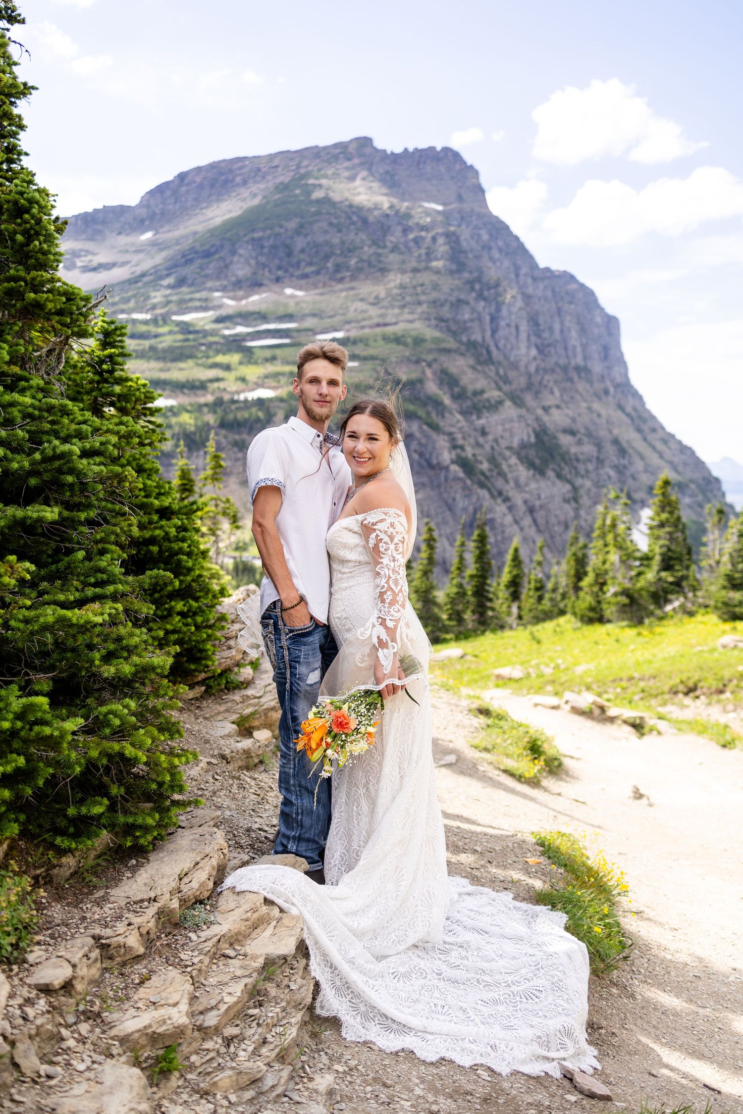 Smiling newlyweds pose on rocky mountain path, bride in lace dress holding colorful bouquet with peaks behind.