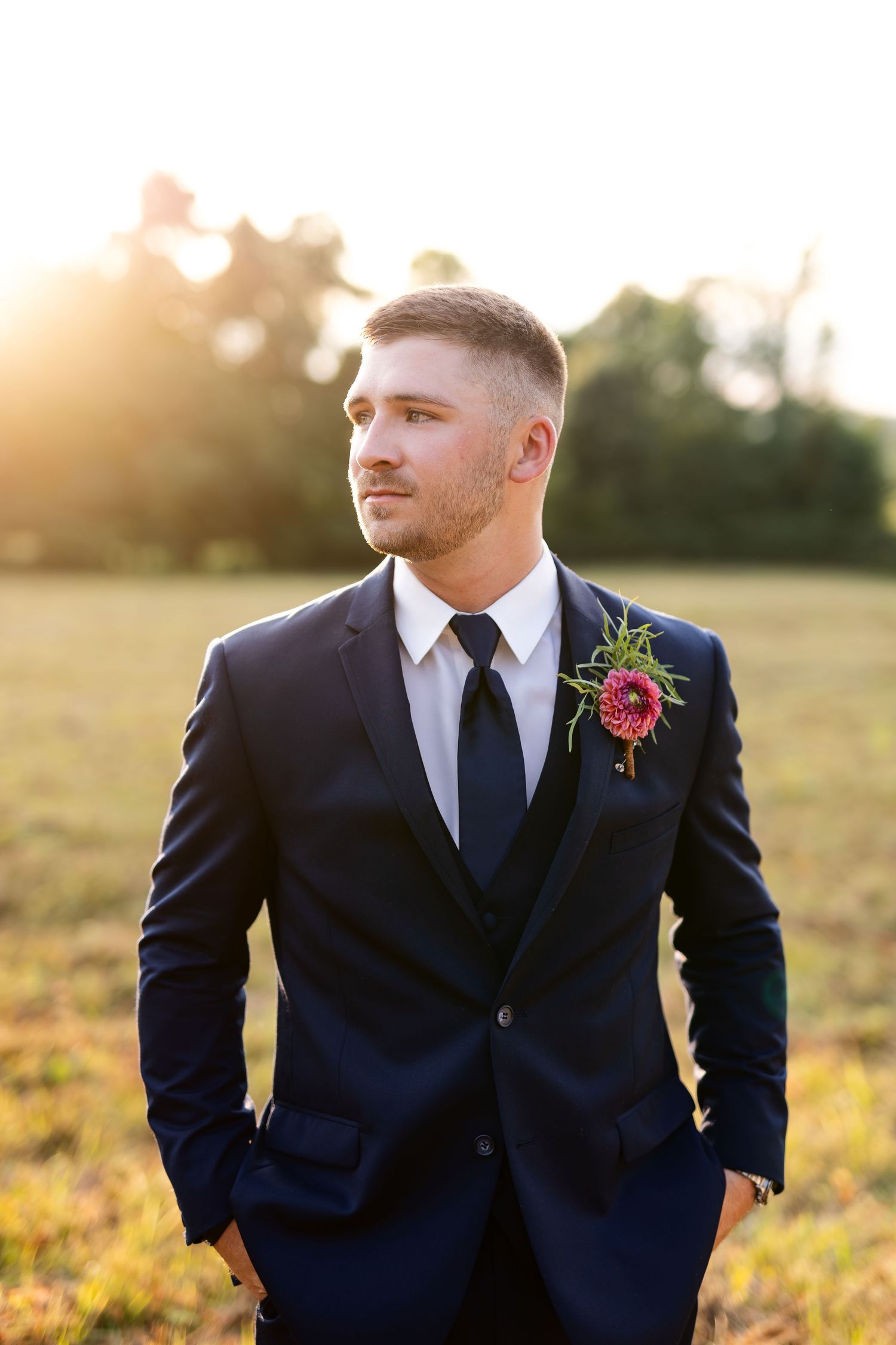 Handsome groom in navy suit with pink boutonniere poses confidently in a golden sunlit outdoor field.