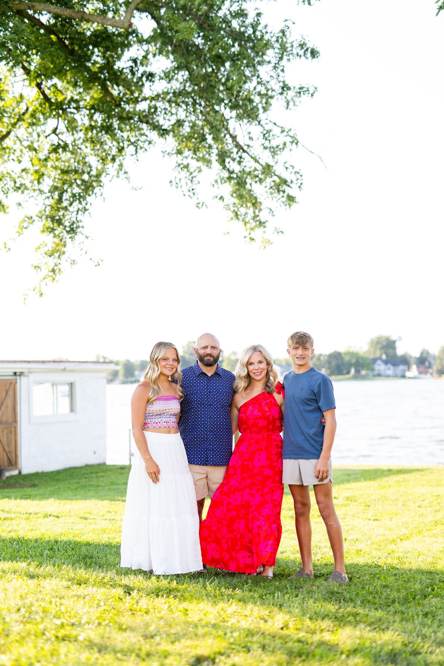 Family of four posing on green lawn near waterfront, wearing colorful summer outfits under a shady tree.