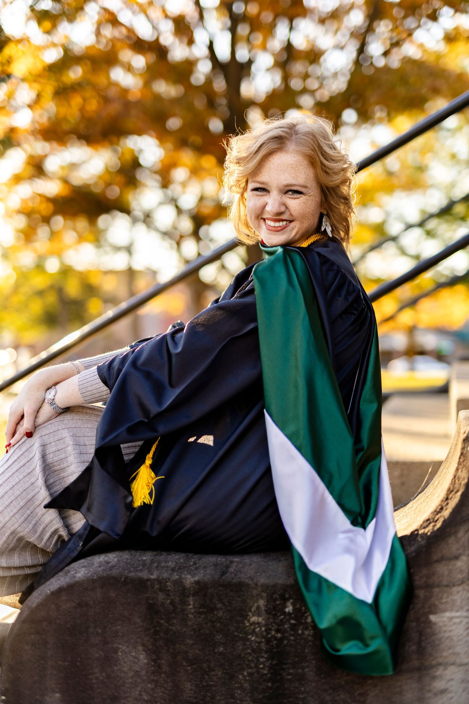 Smiling blonde woman in graduation gown with green hood sitting on stone steps among autumn foliage.