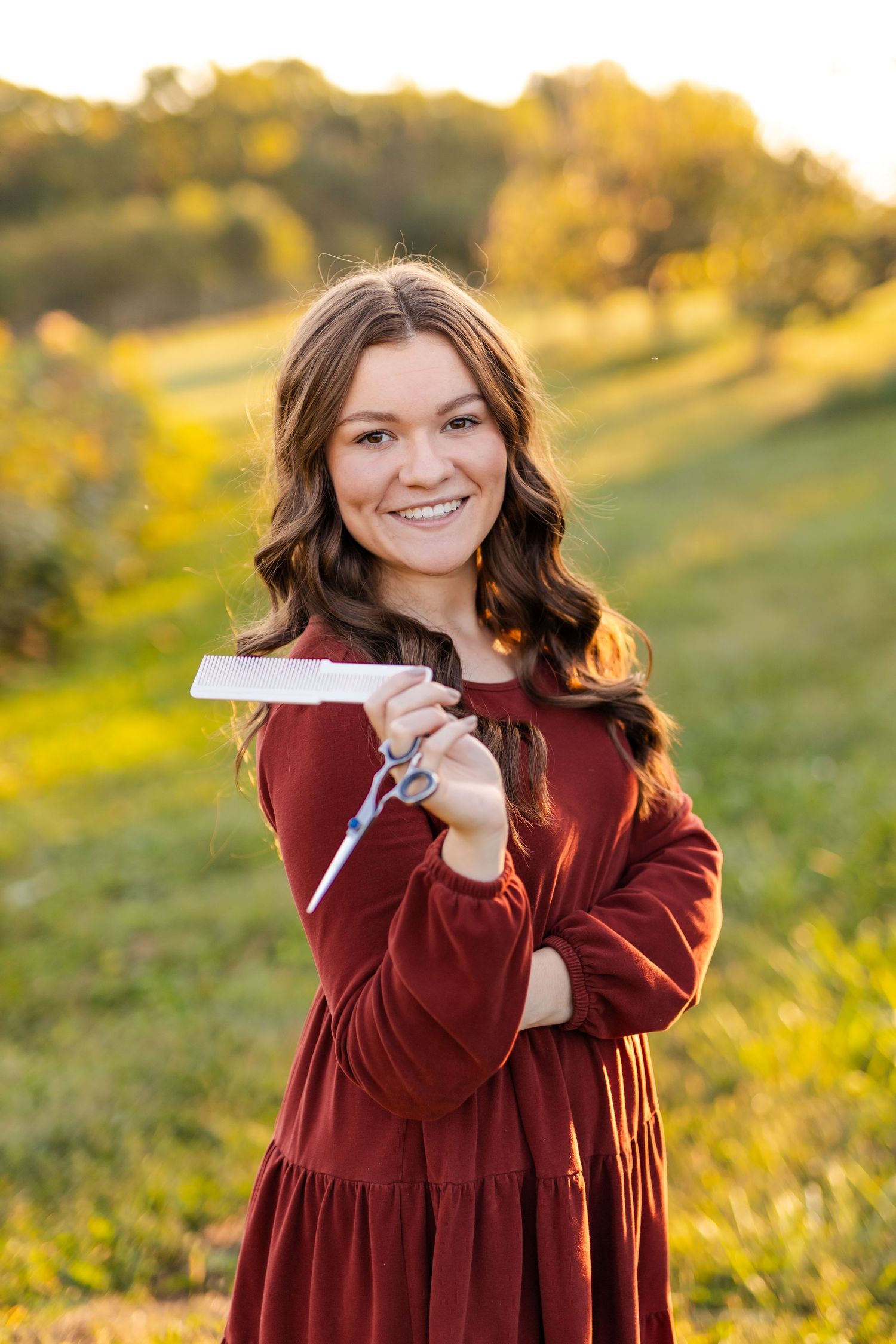 Smiling young woman in burgundy dress holding scissors and comb outdoors in golden sunlight among green hills.