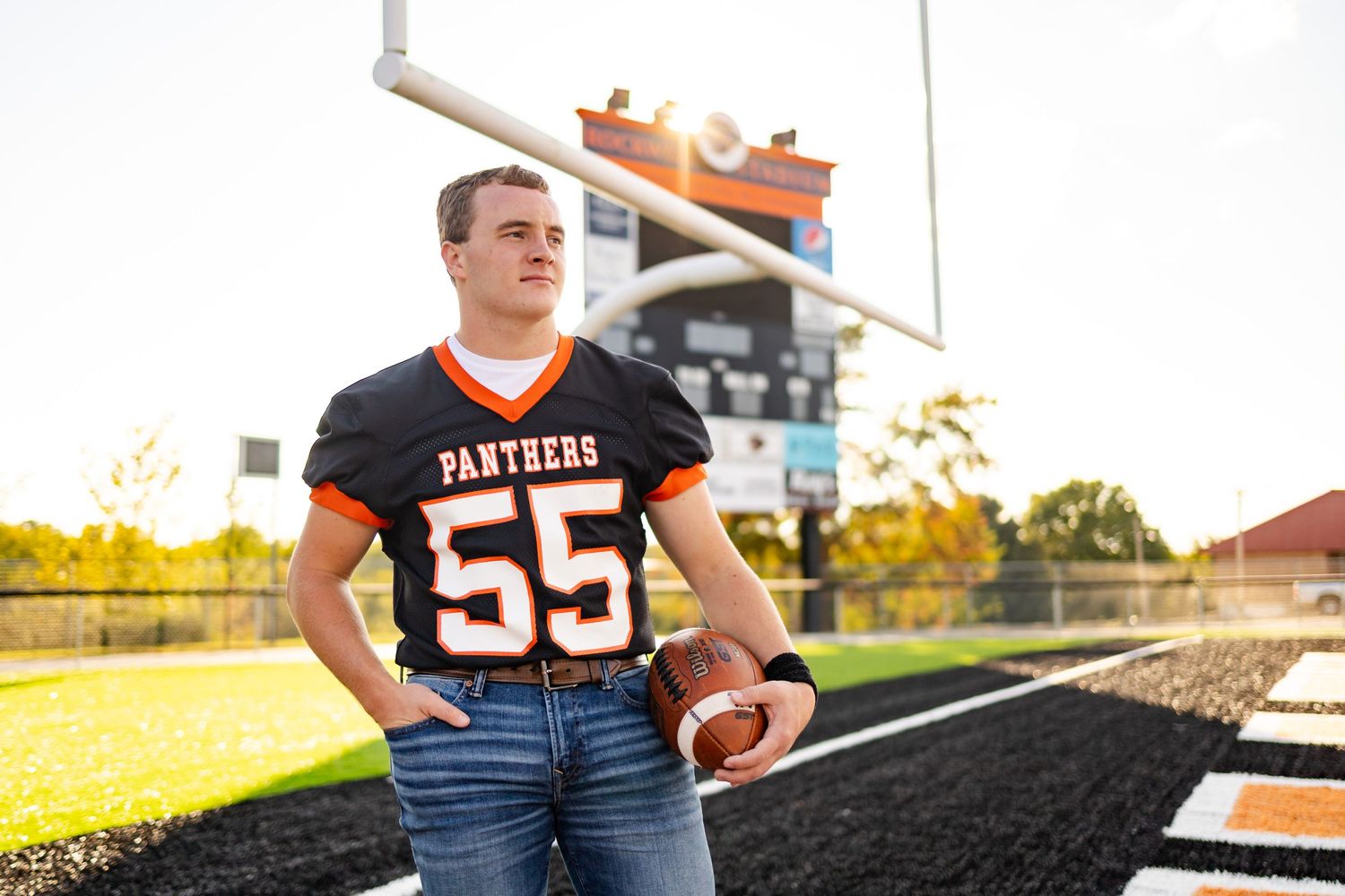 High school football player in Panthers jersey number 55 holding a football on the field during senior photos.