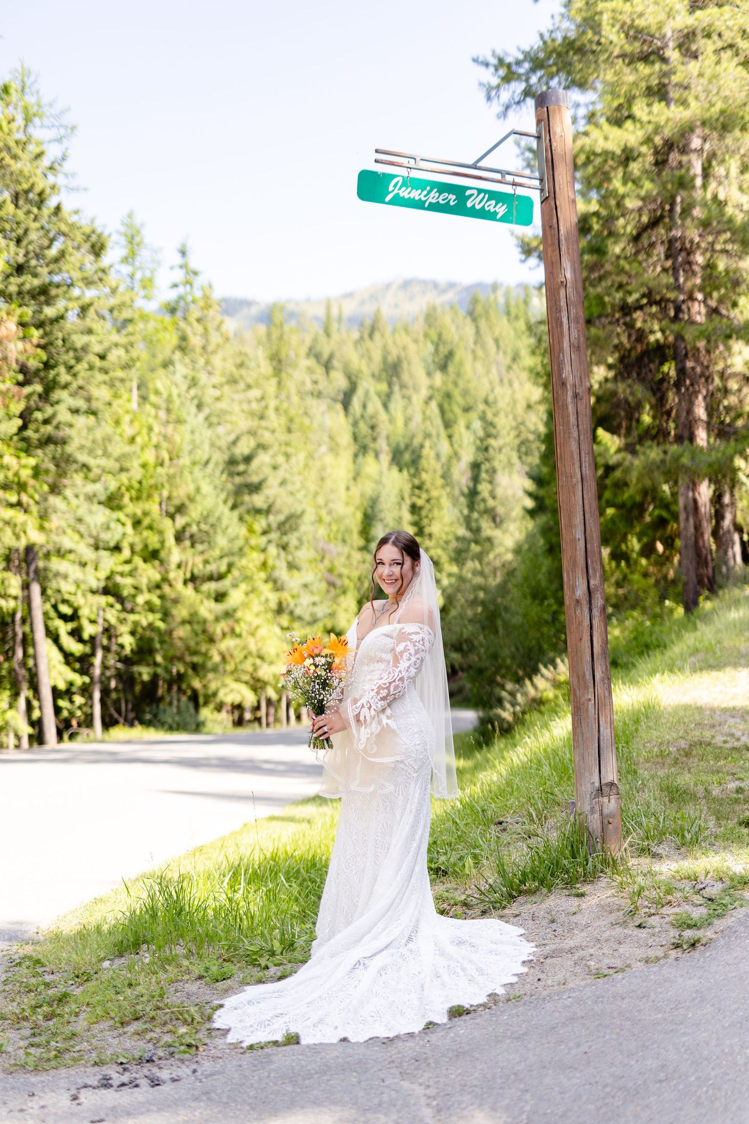 Smiling bride in lace gown and veil poses by Juniper Way sign surrounded by lush mountain forest.