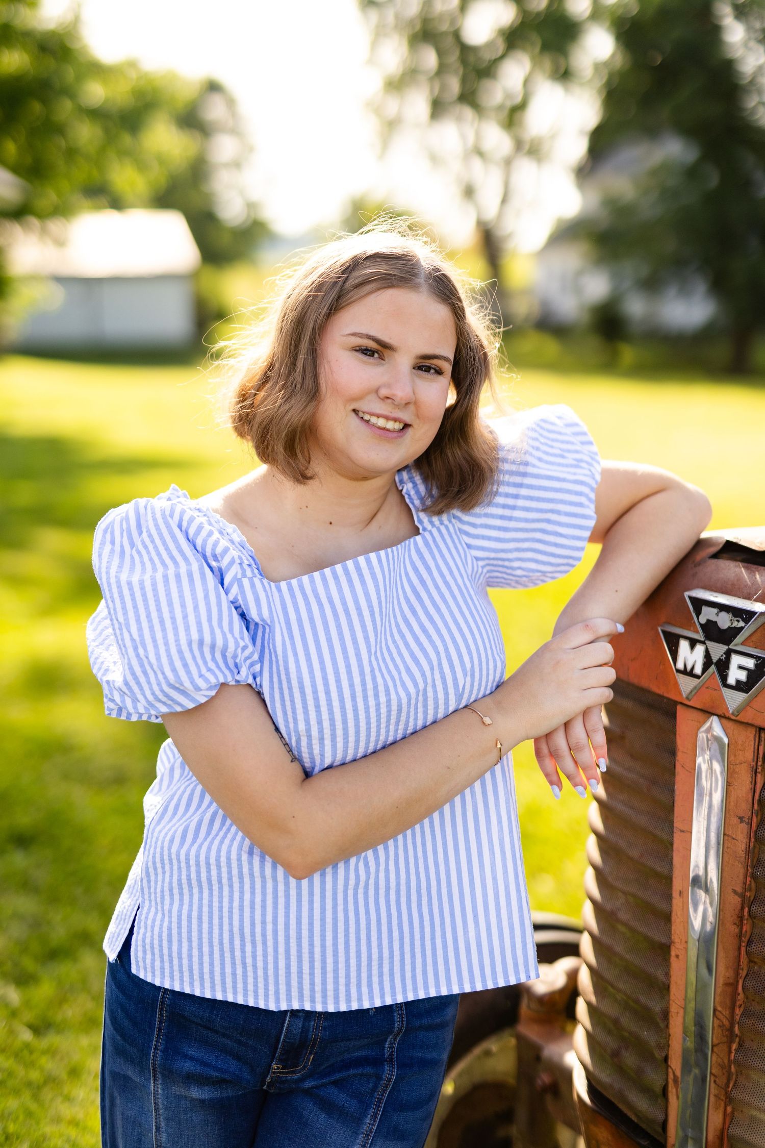 Smiling teenage girl in striped off-shoulder top leaning on vintage tractor in golden hour light.