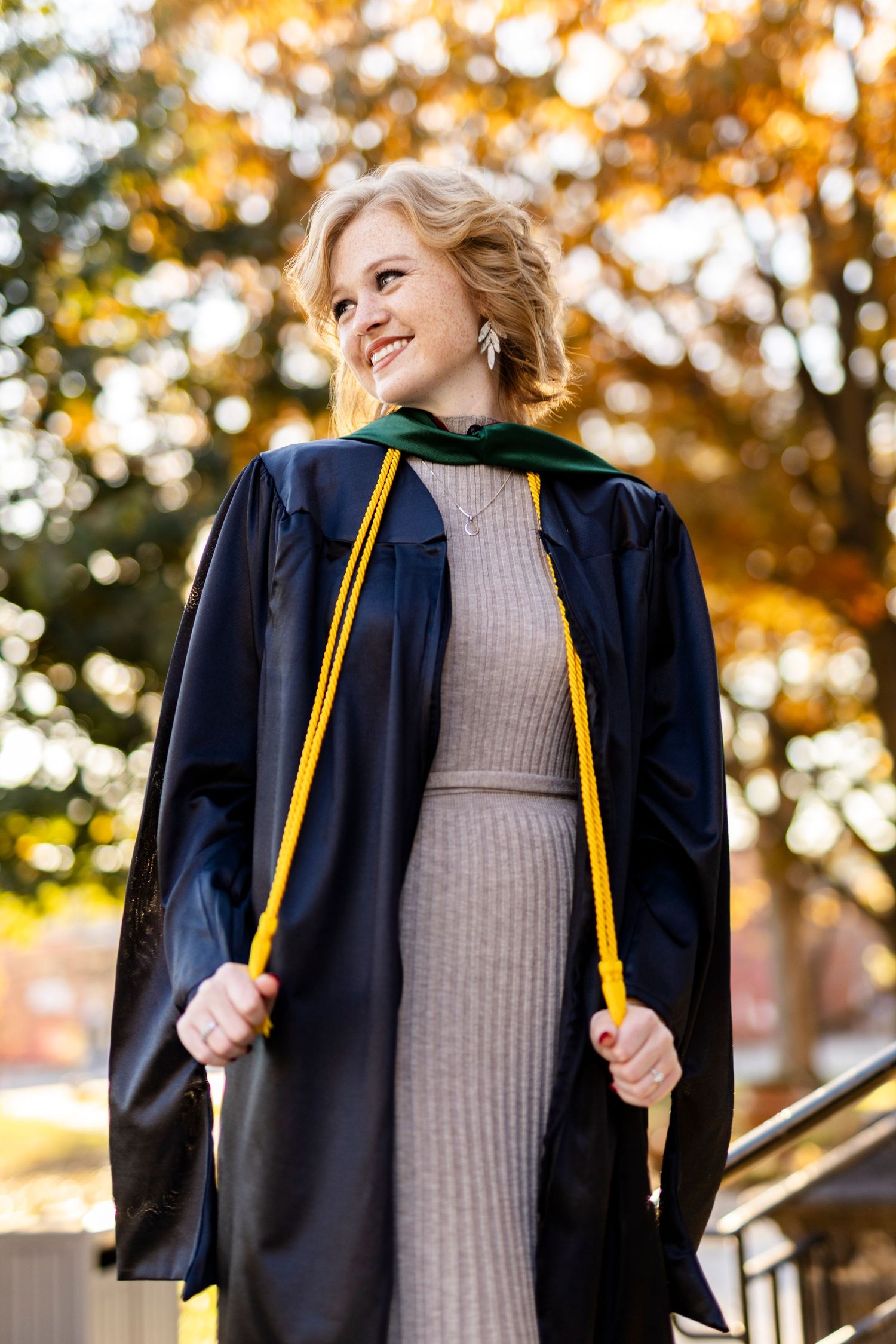 Smiling blonde woman in black graduation gown with green hood and gold honor cords outdoors in autumn.