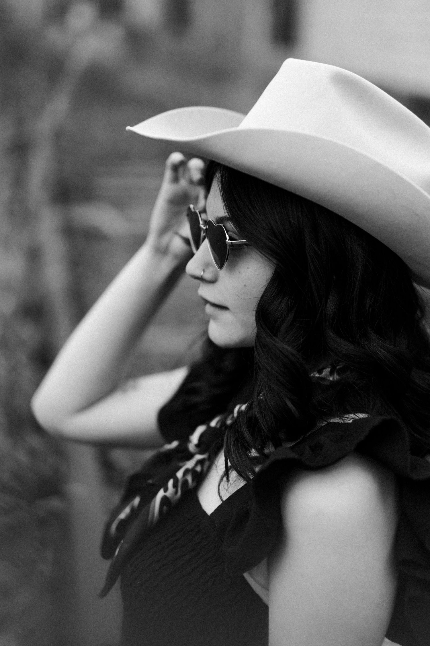 Black and white portrait of a woman in a cowboy hat and sunglasses, holding her hat brim outdoors.