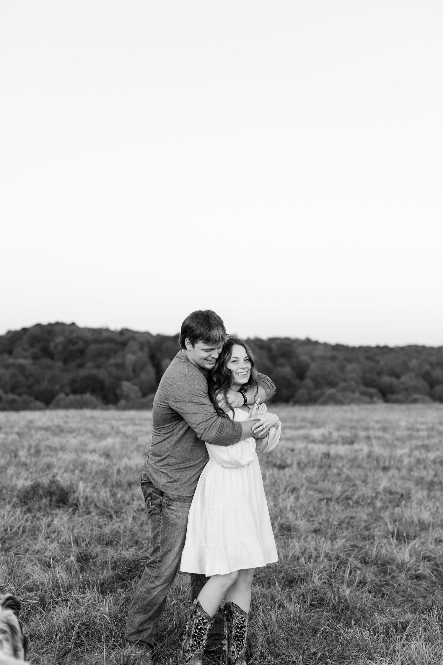 Black and white photo of a couple embracing in an open field, the woman smiling in a white dress.
