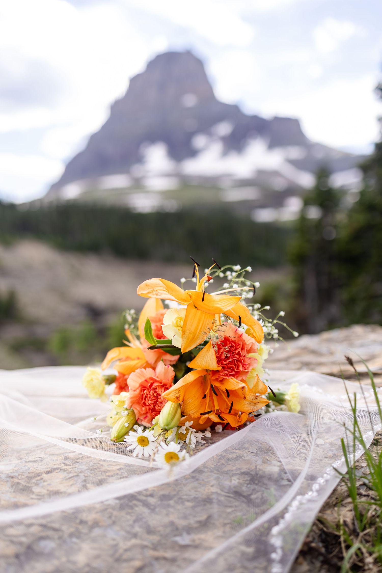 Vibrant orange lily and carnation wedding bouquet resting on rock with snow-capped mountain backdrop.