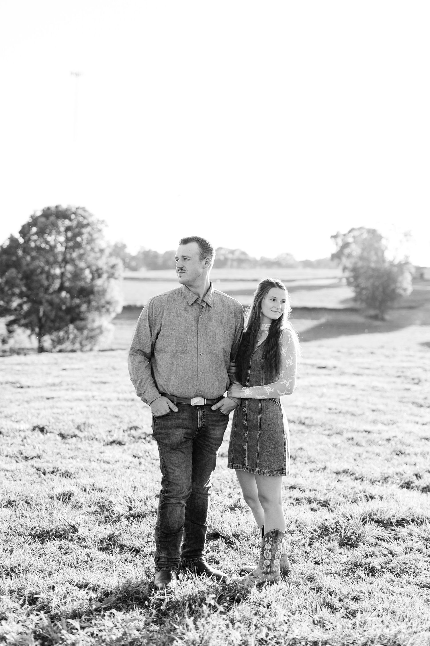 Black and white photo of a couple standing in a field, woman in dress and boots, man in jeans and shirt.