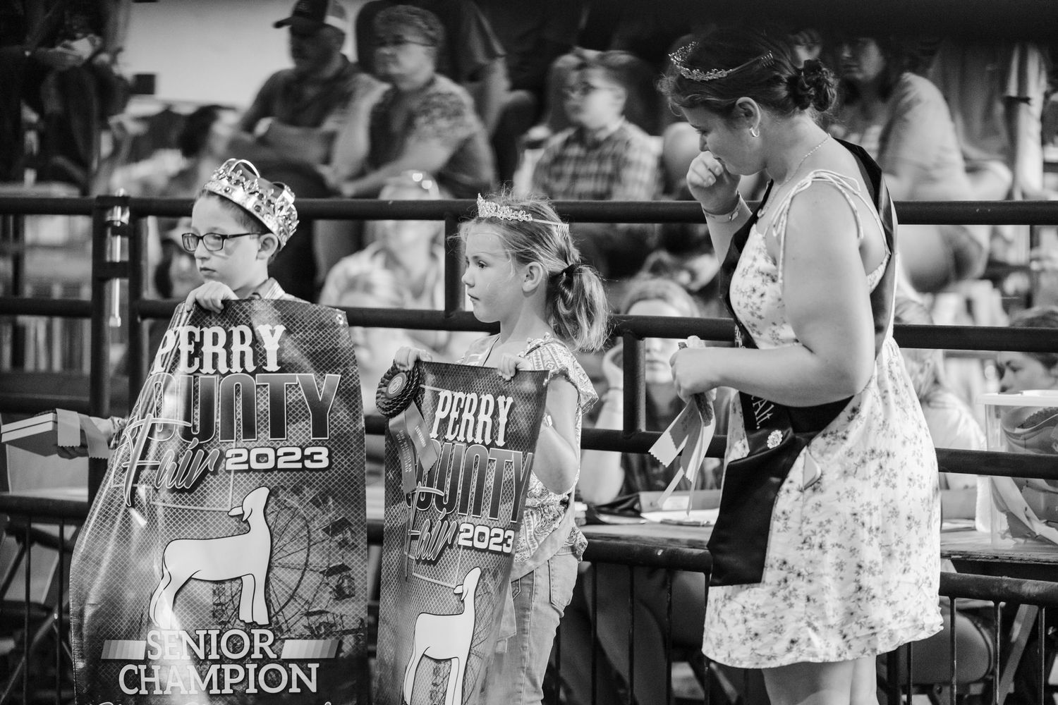 Two young girls wearing Perry July 2023 Junior Champion ribbons stand with a woman at a fair.