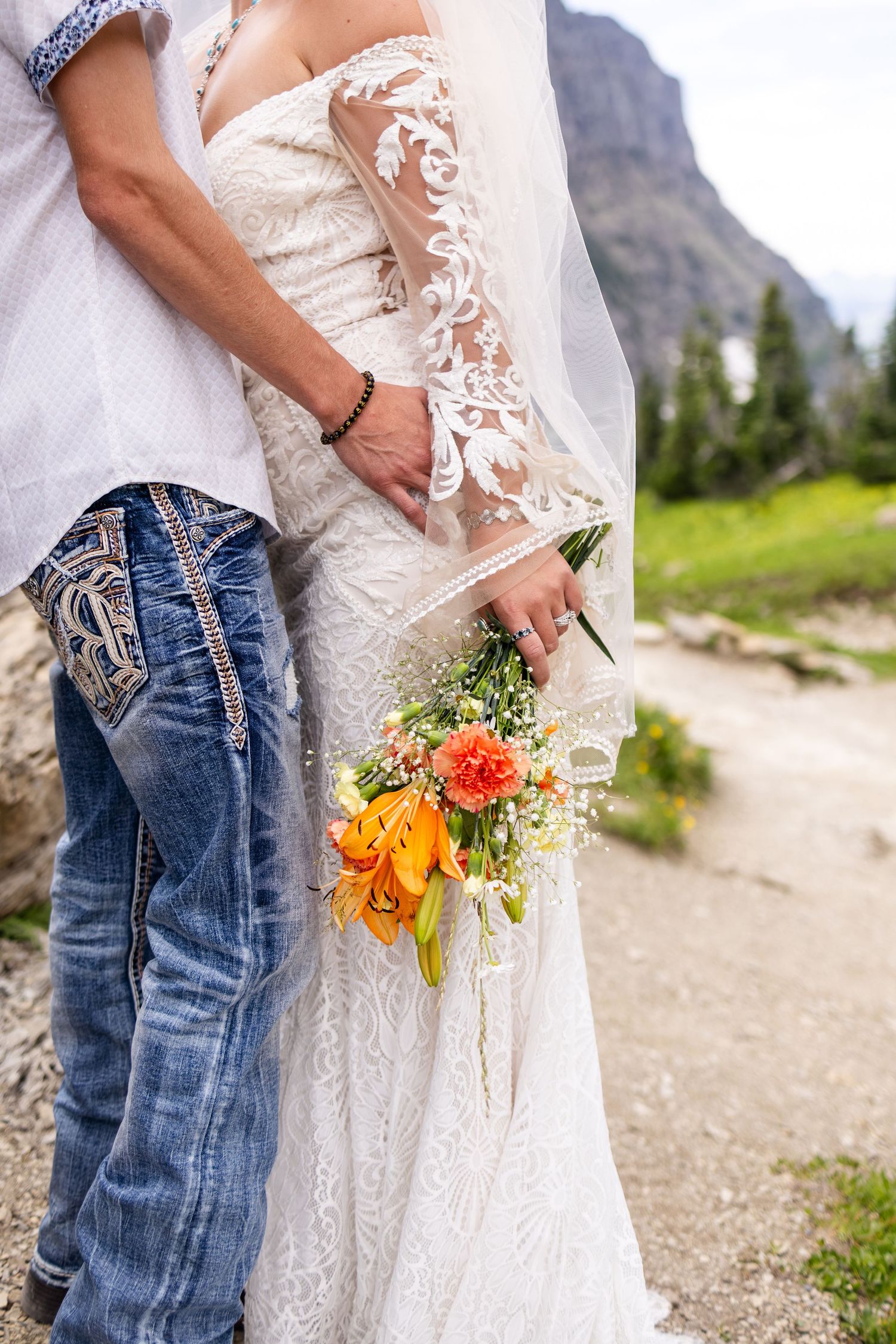 Close-up of bride in lace wedding dress holding vibrant orange and yellow bouquet with groom in jeans.