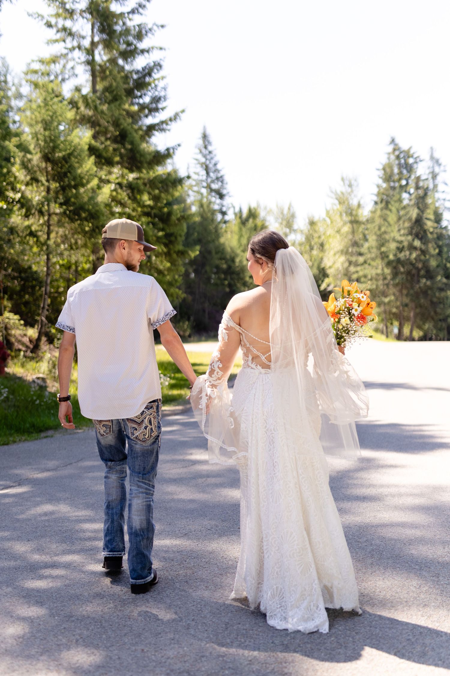 Bride in lace gown holding bouquet walks hand-in-hand with groom in jeans and cap on a sunny tree-lined path.