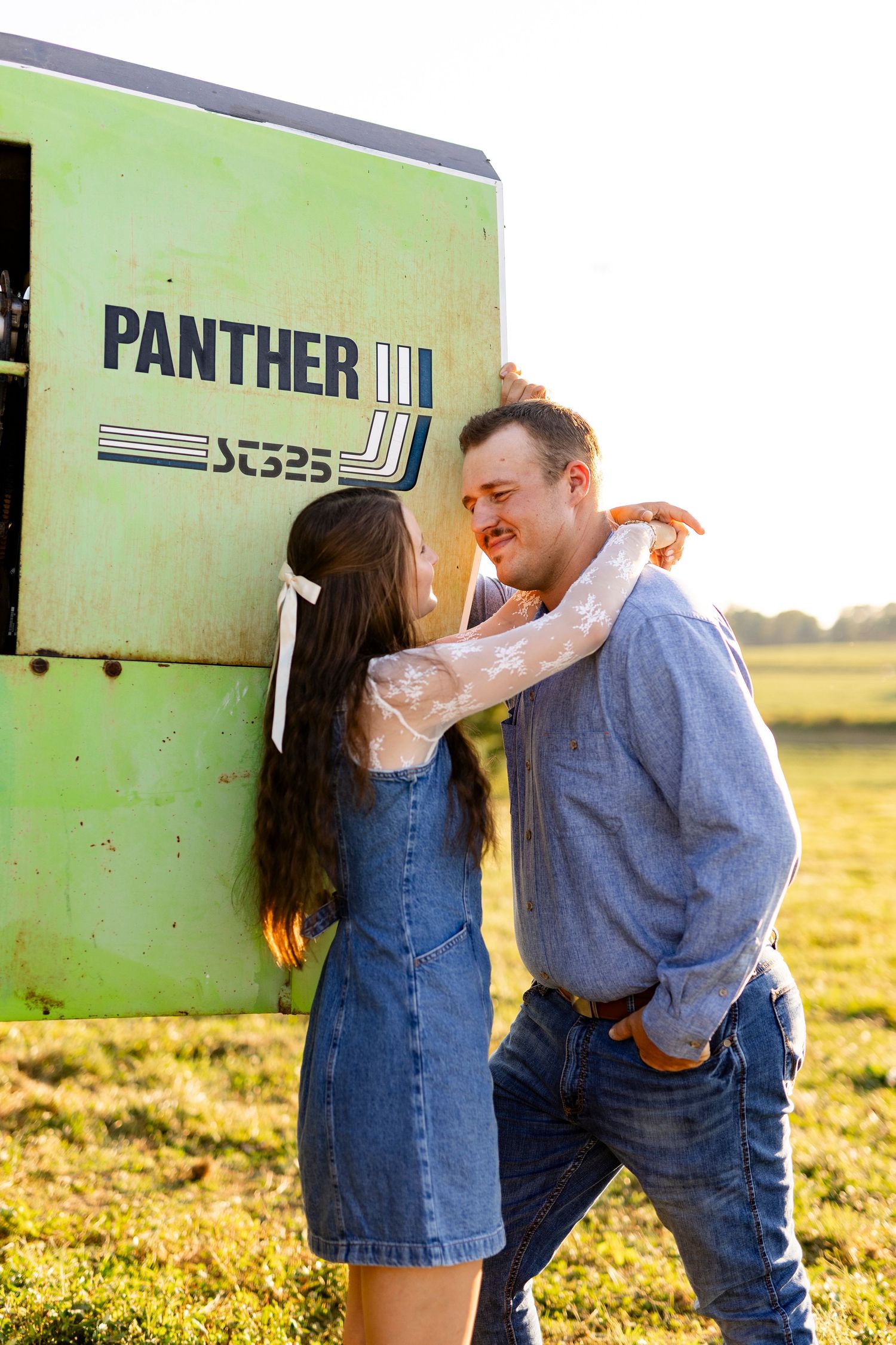 Couple embracing and smiling by a green Panther III ST325 machine in a sunny field, wearing denim outfits.
