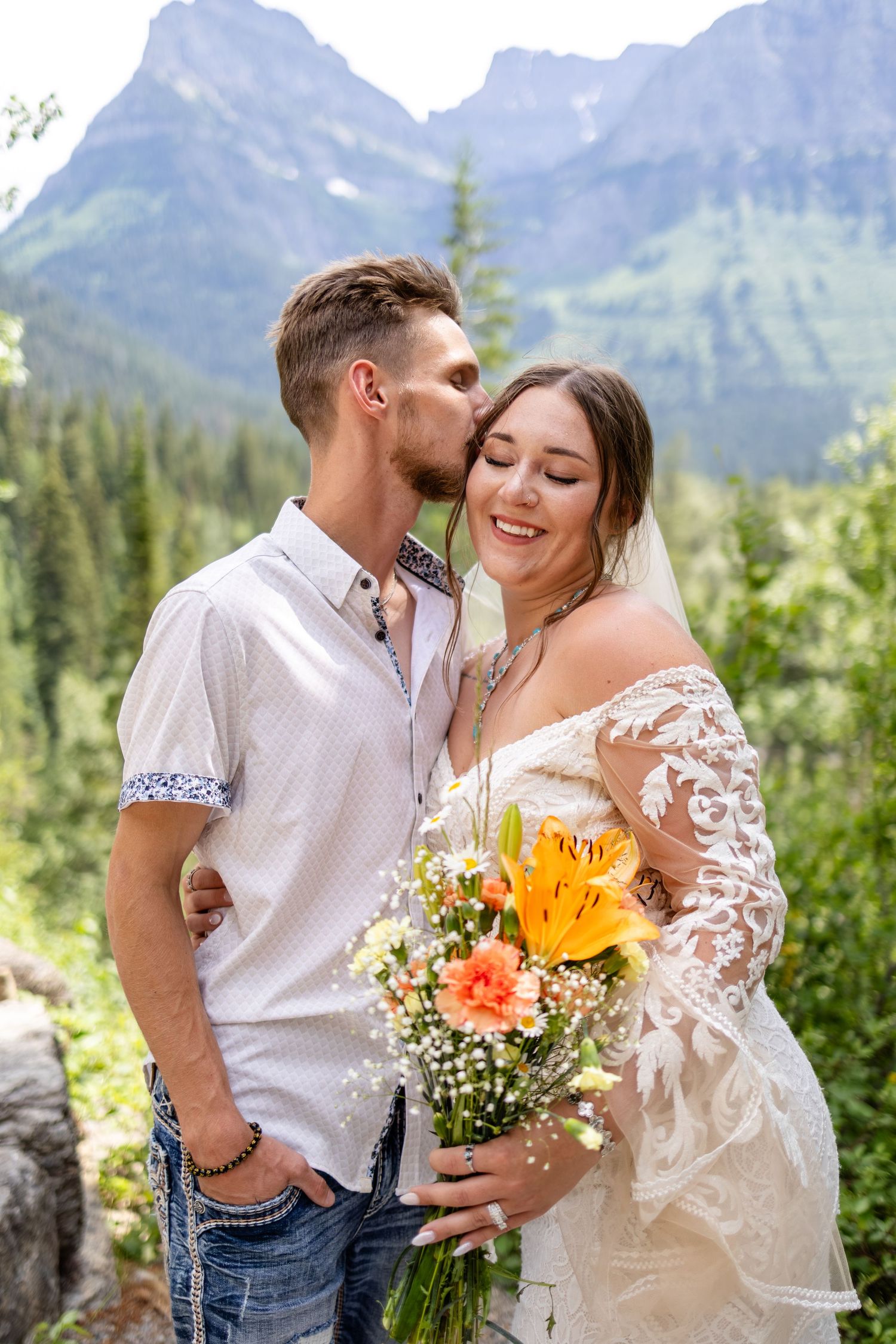 Groom kisses bride holding yellow lily bouquet with mountain forest background.