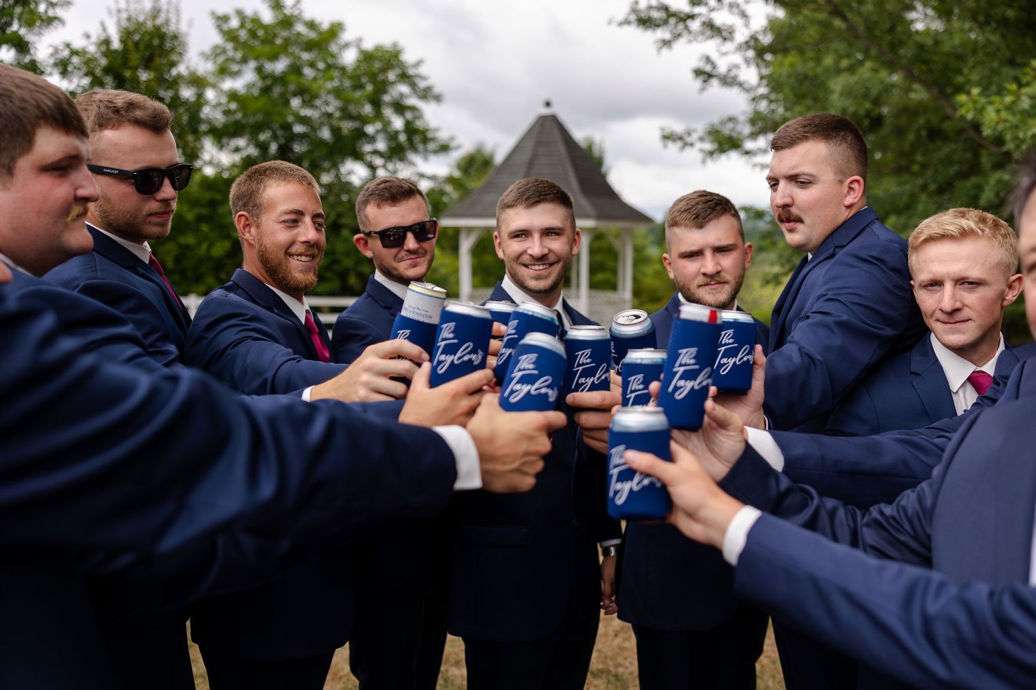 Groomsmen in navy suits toast with canned beverages outdoors near a white gazebo before the wedding.