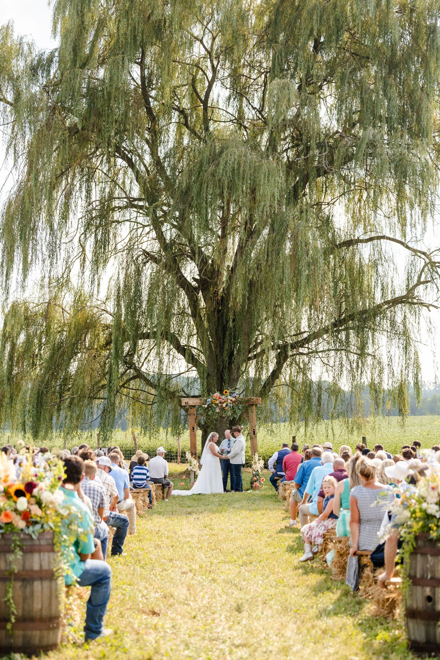 Guests seated along a rustic aisle with barrel flower arrangements watch a couple wed beneath a large weeping willow tree.