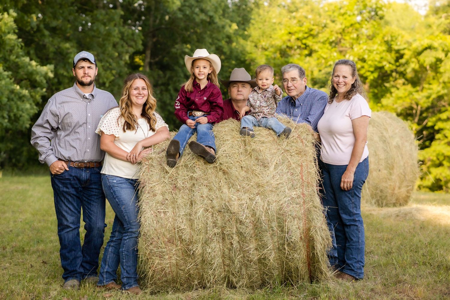 A family of seven poses together around a large round hay bale in a green field.