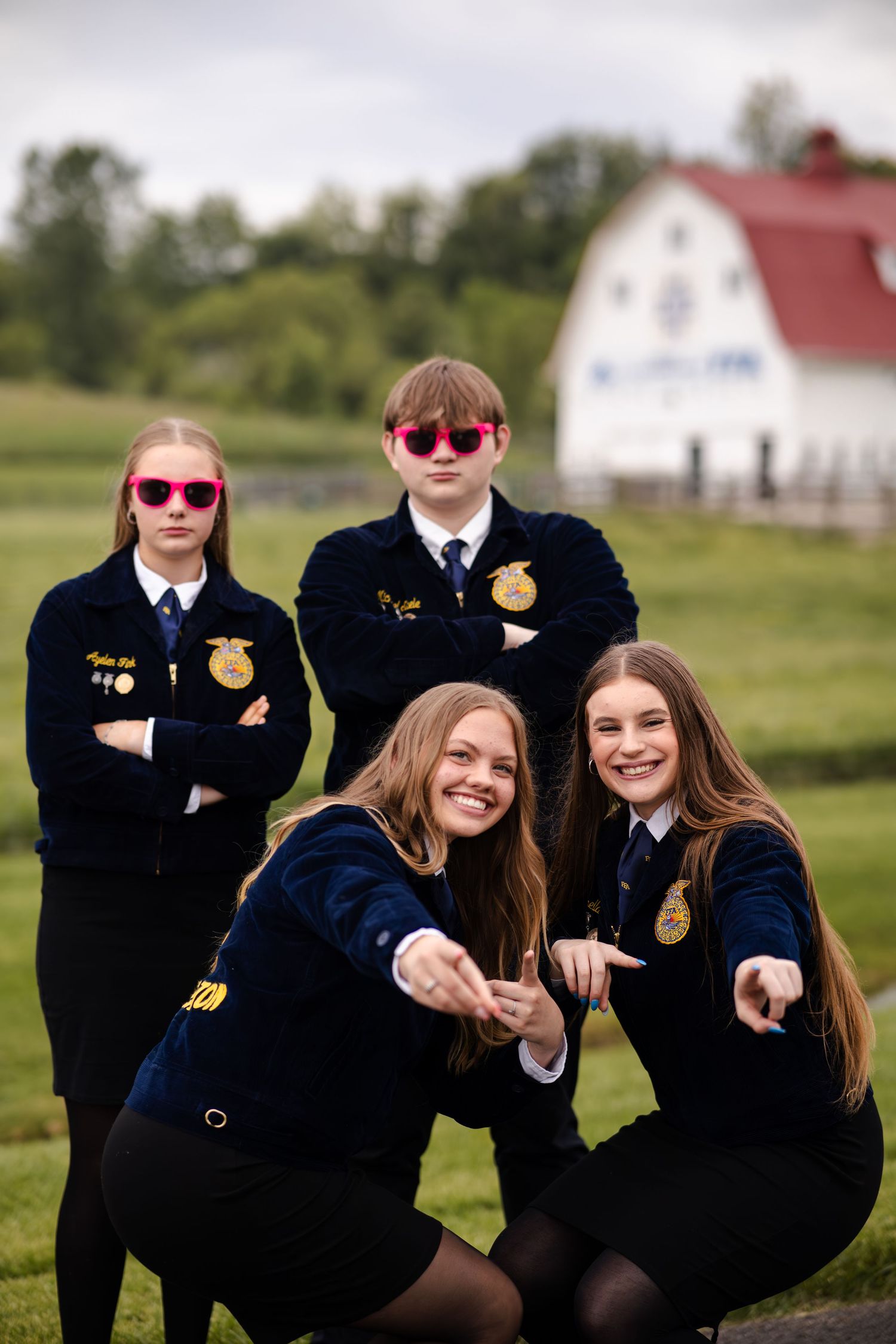 Four FFA students posing playfully with pink sunglasses near a white barn outdoors.